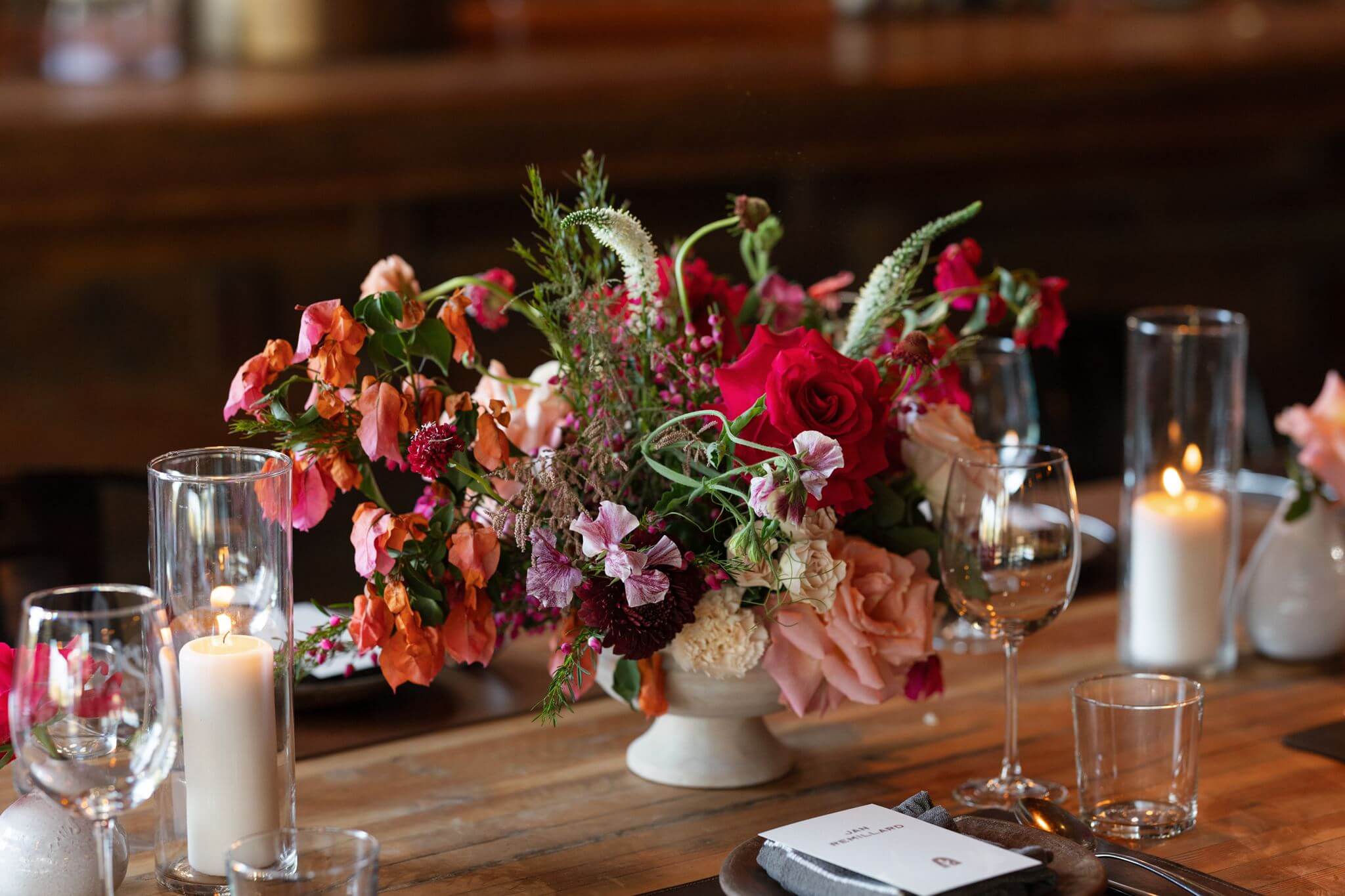 Colorful floral wedding centerpiece with pink and red flowers surrounded by candles and glassware.
