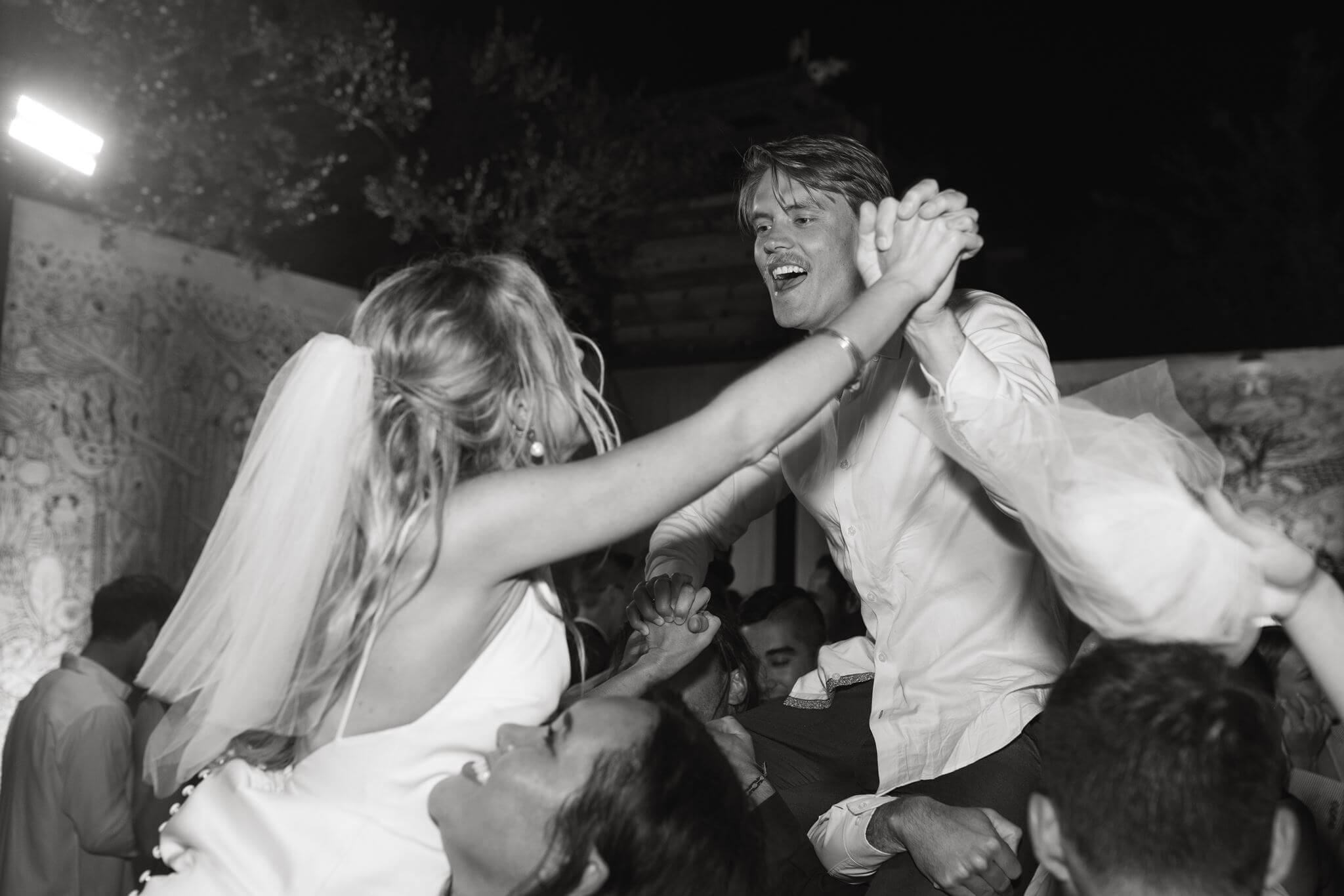 Black-and-white photo of the bride and groom dancing above the crowd during their nighttime reception.