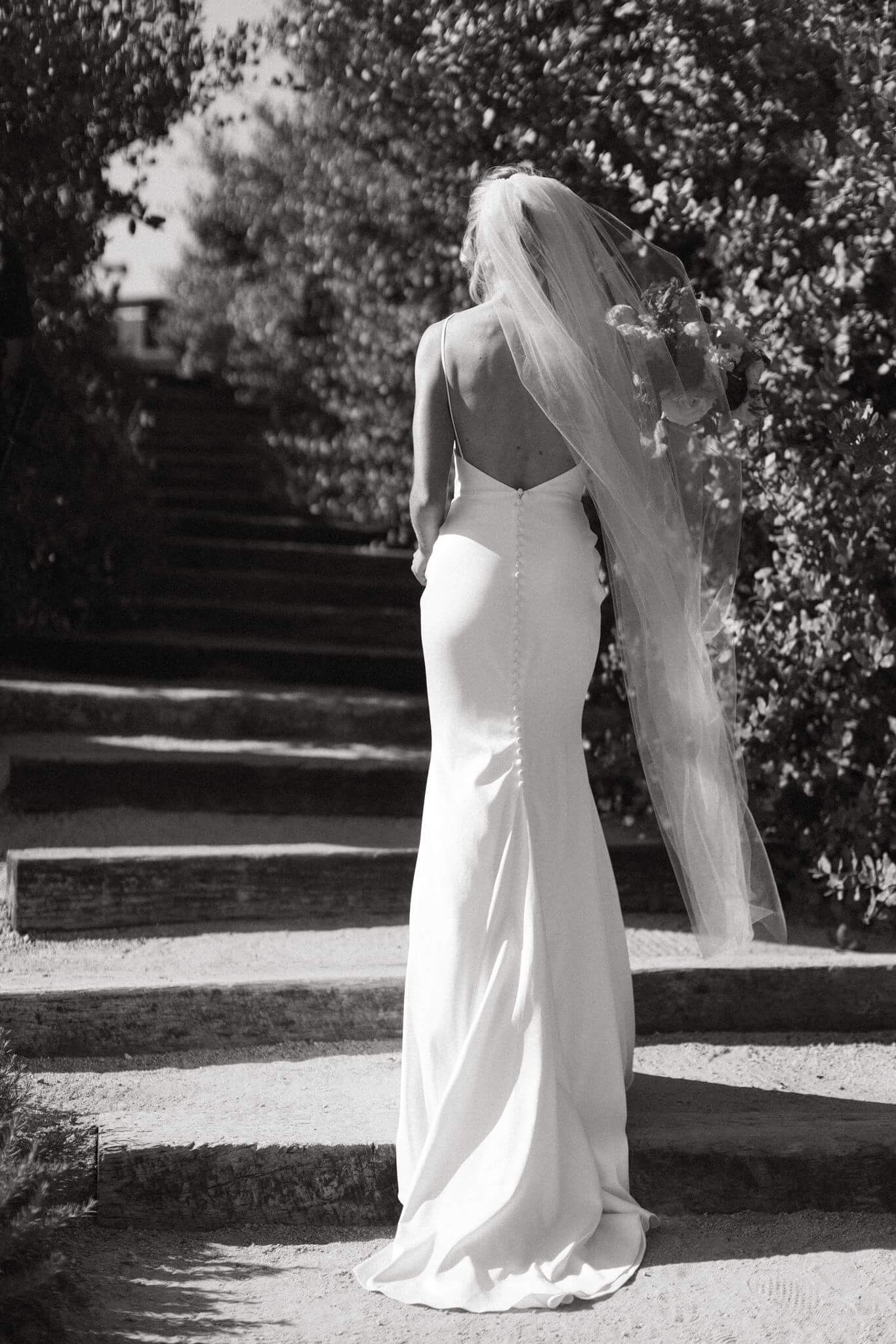 Black-and-white portrait of the bride walking up outdoor steps with her long veil trailing behind her.