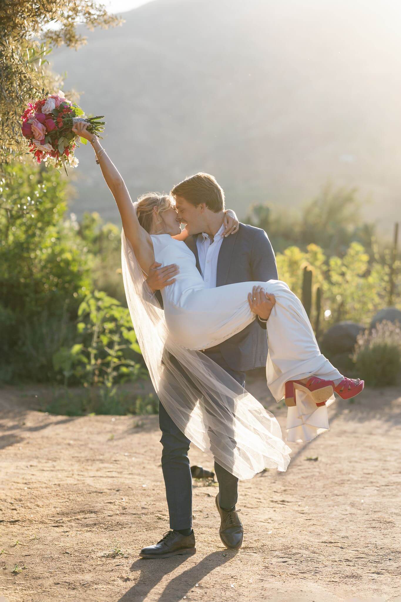Groom carrying the bride as she raises her bouquet during playful sunset wedding portraits.