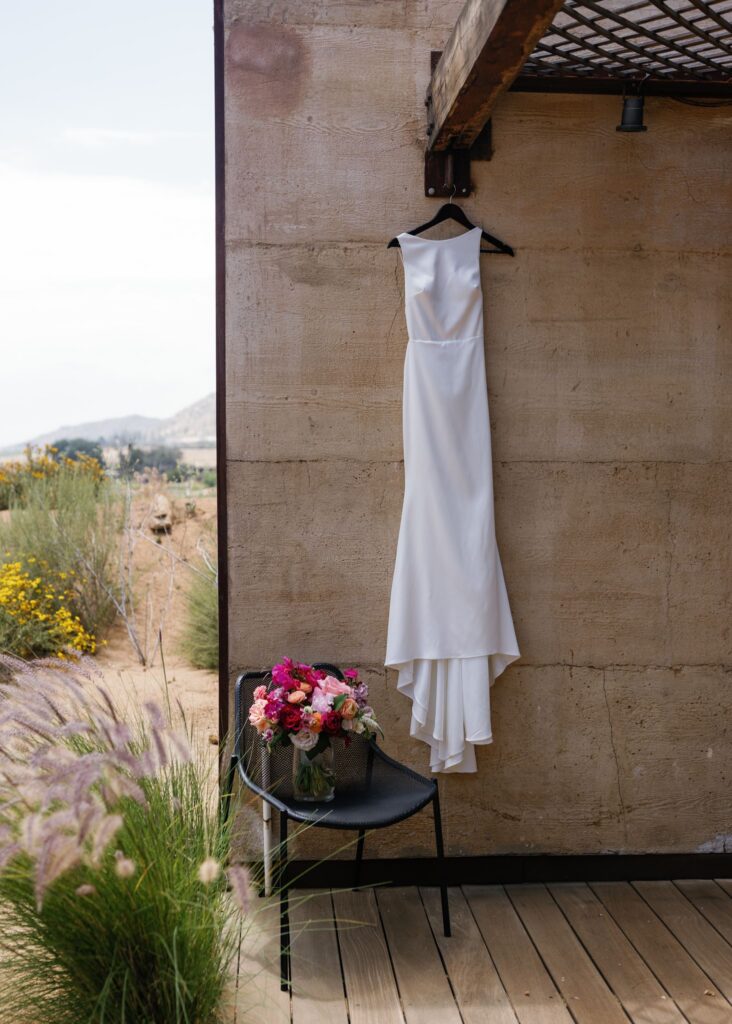 Bride’s white wedding dress hanging outside a modern concrete suite beside her colorful bouquet.