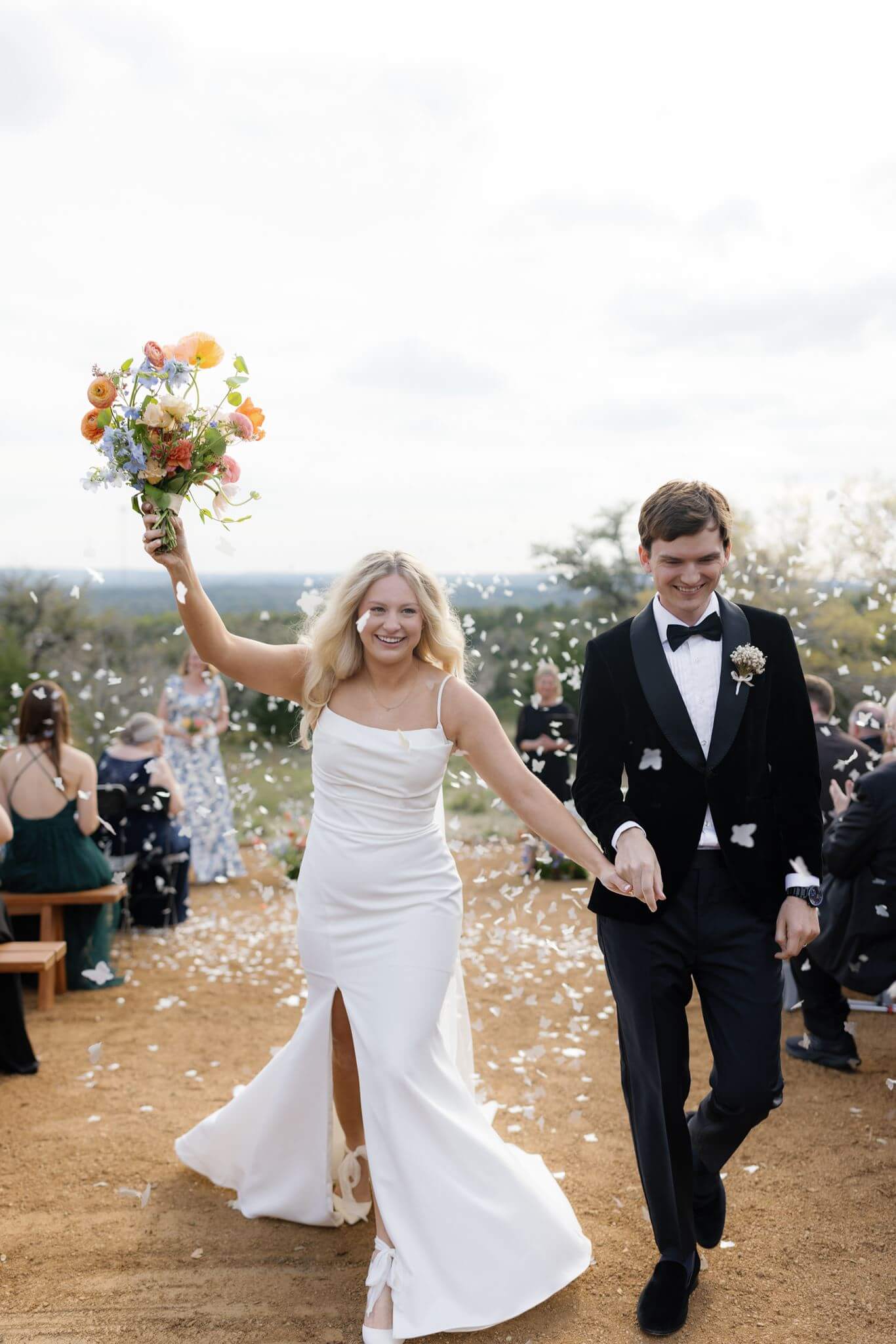 Bride raising her bouquet in celebration while walking hand in hand with groom through confetti