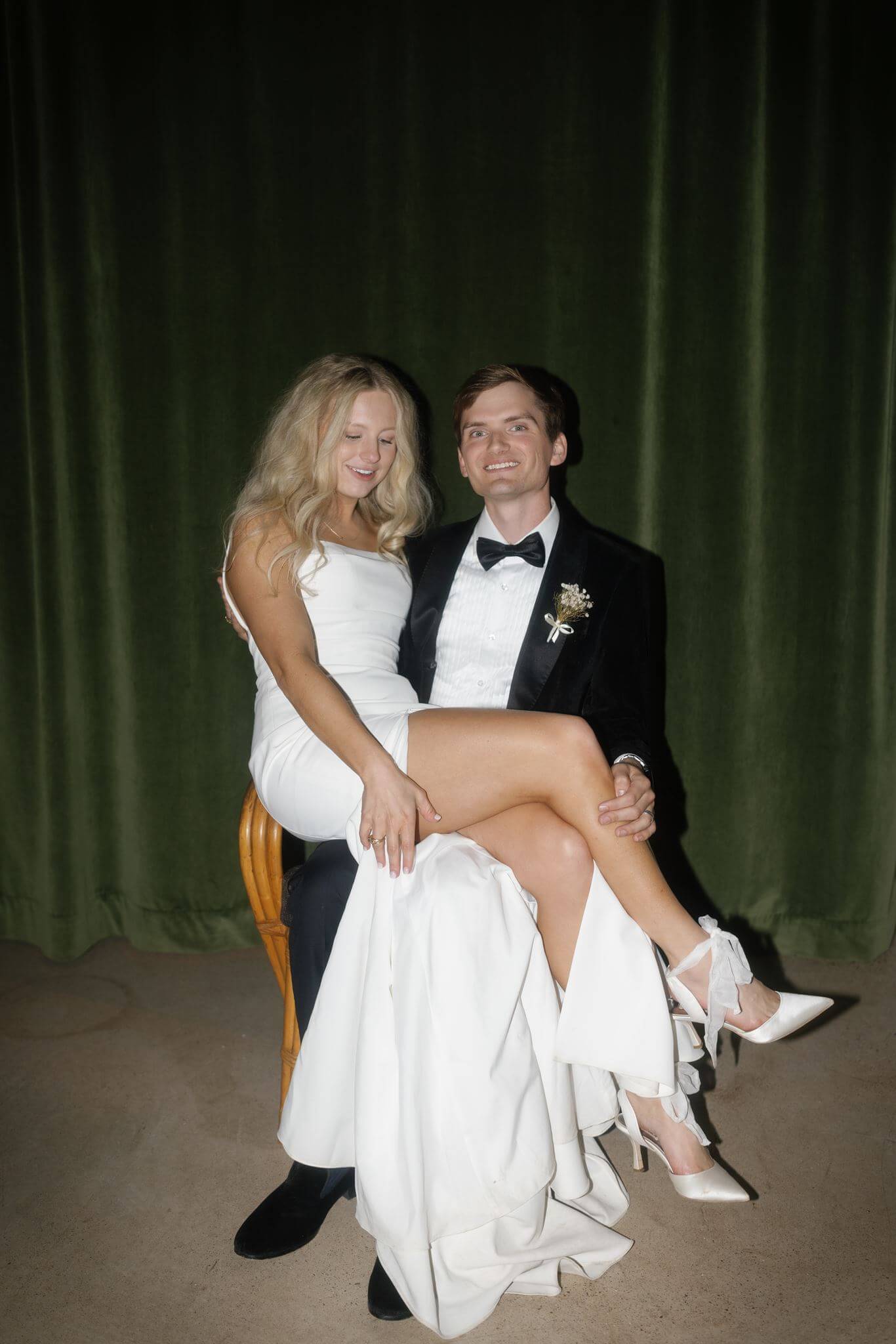 Bride sitting on groom's lap with her leg crossed, both smiling at the camera