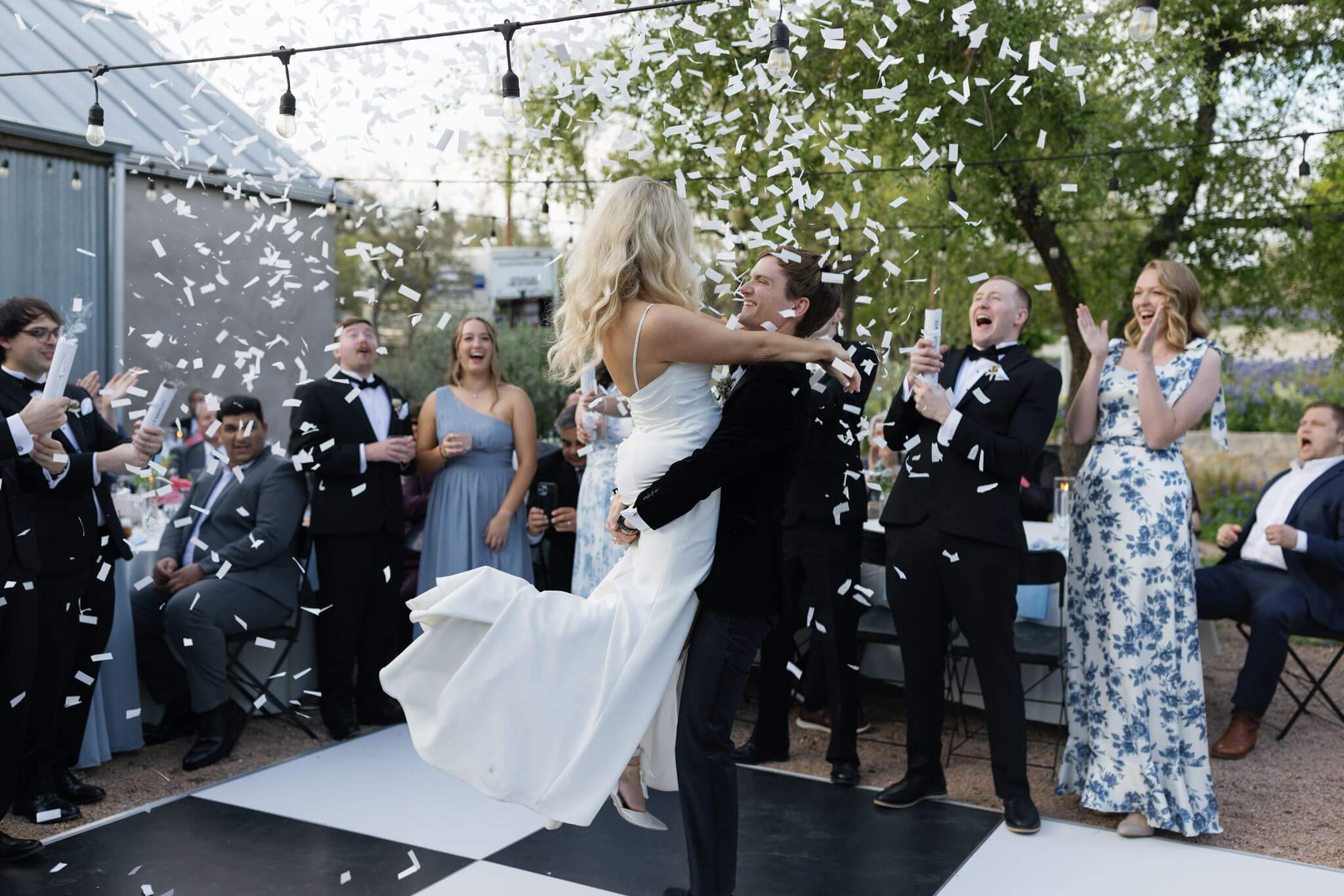 Wide shot of bride and groom kissing amid a shower of confetti as wedding guests cheer around them