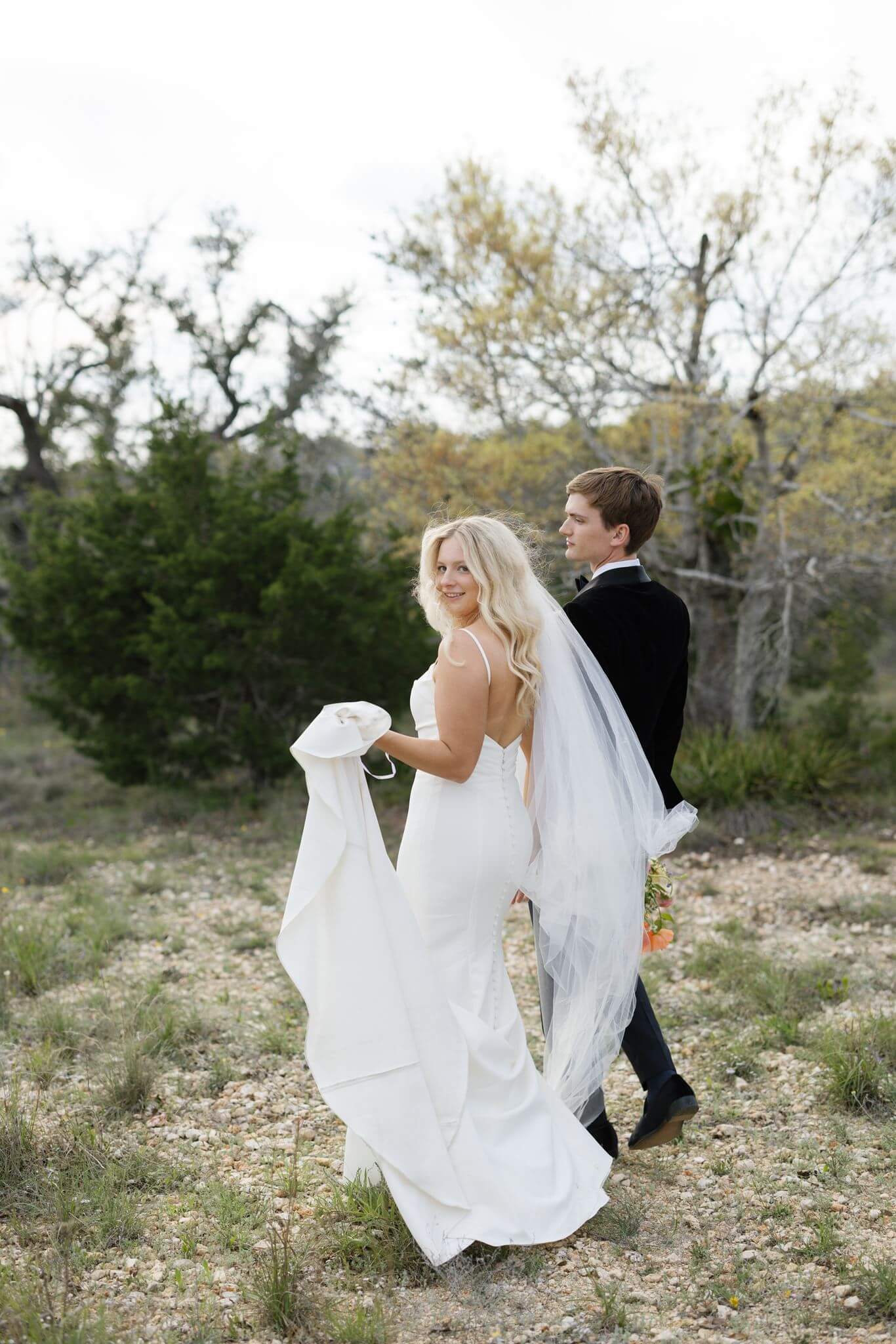 Bride and groom walking hand in hand across a wildflower field, her veil trailing in the wind