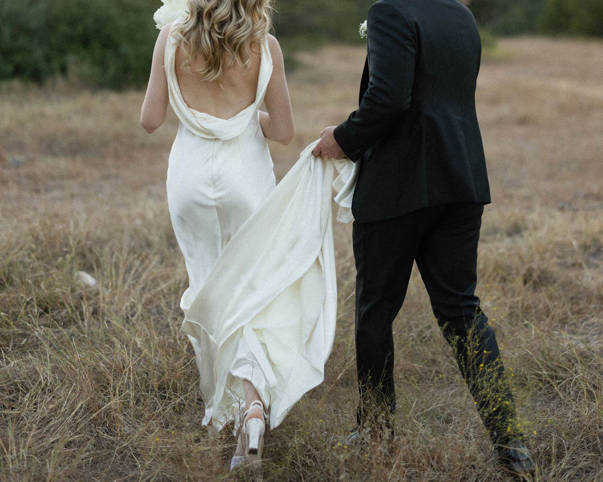 Bride and groom walk through a grassy field from behind as the groom carries the train of her wedding dress.