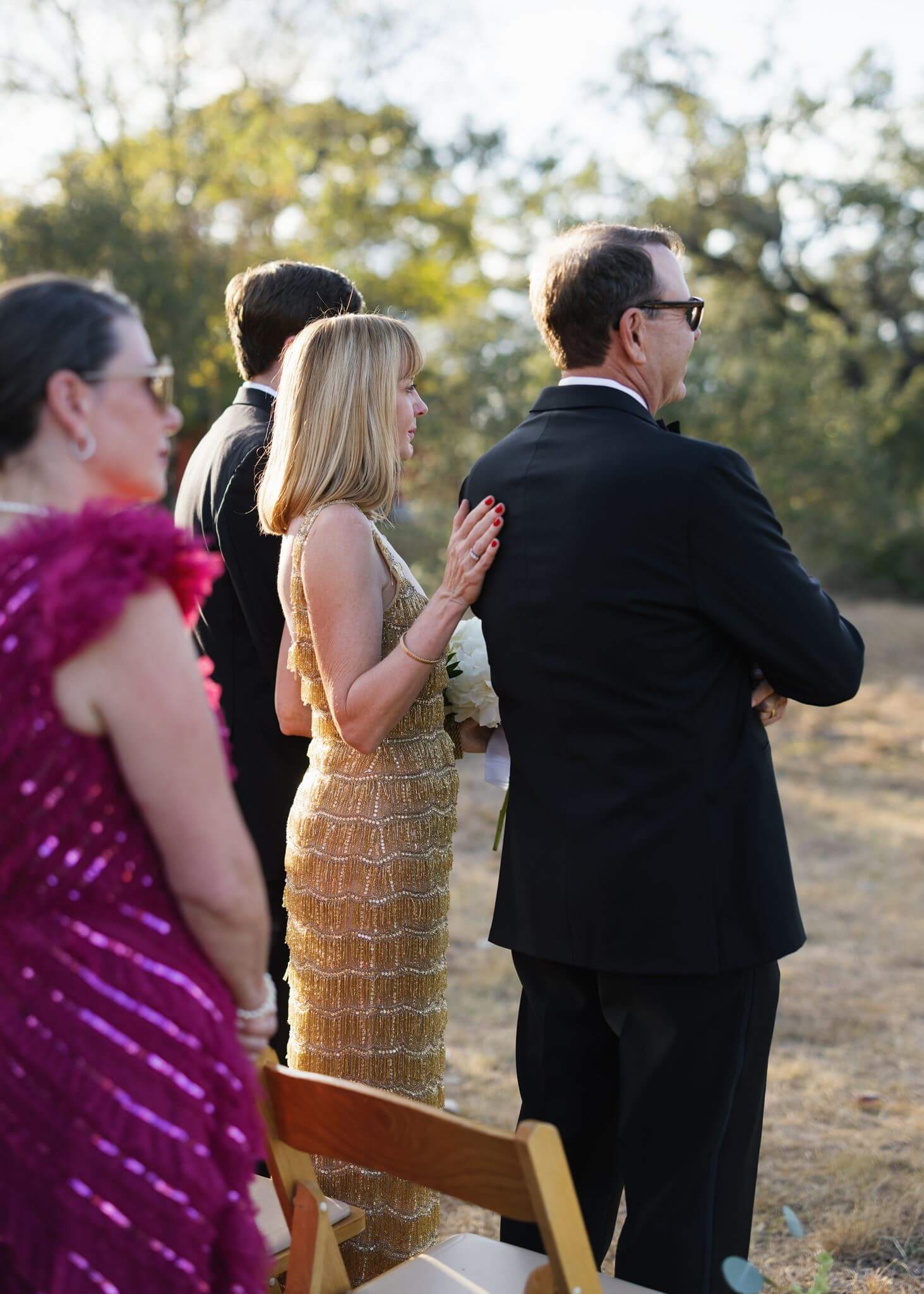 Wedding guests watch the outdoor ceremony from wooden chairs, including a woman wearing a gold sequined dress.