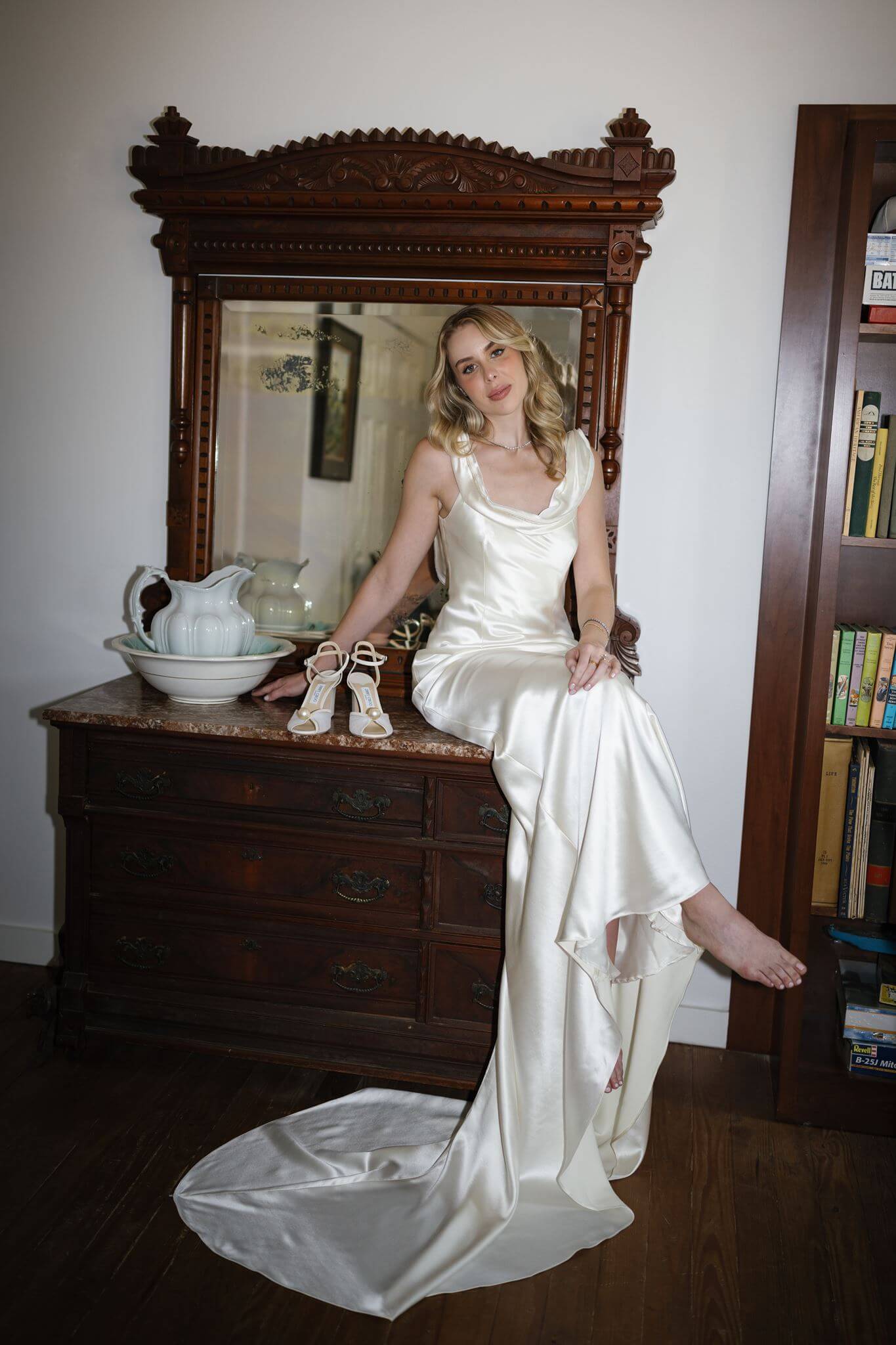 Bride poses beside an antique dresser and ornate mirror indoors while lifting the skirt of her satin wedding dress.