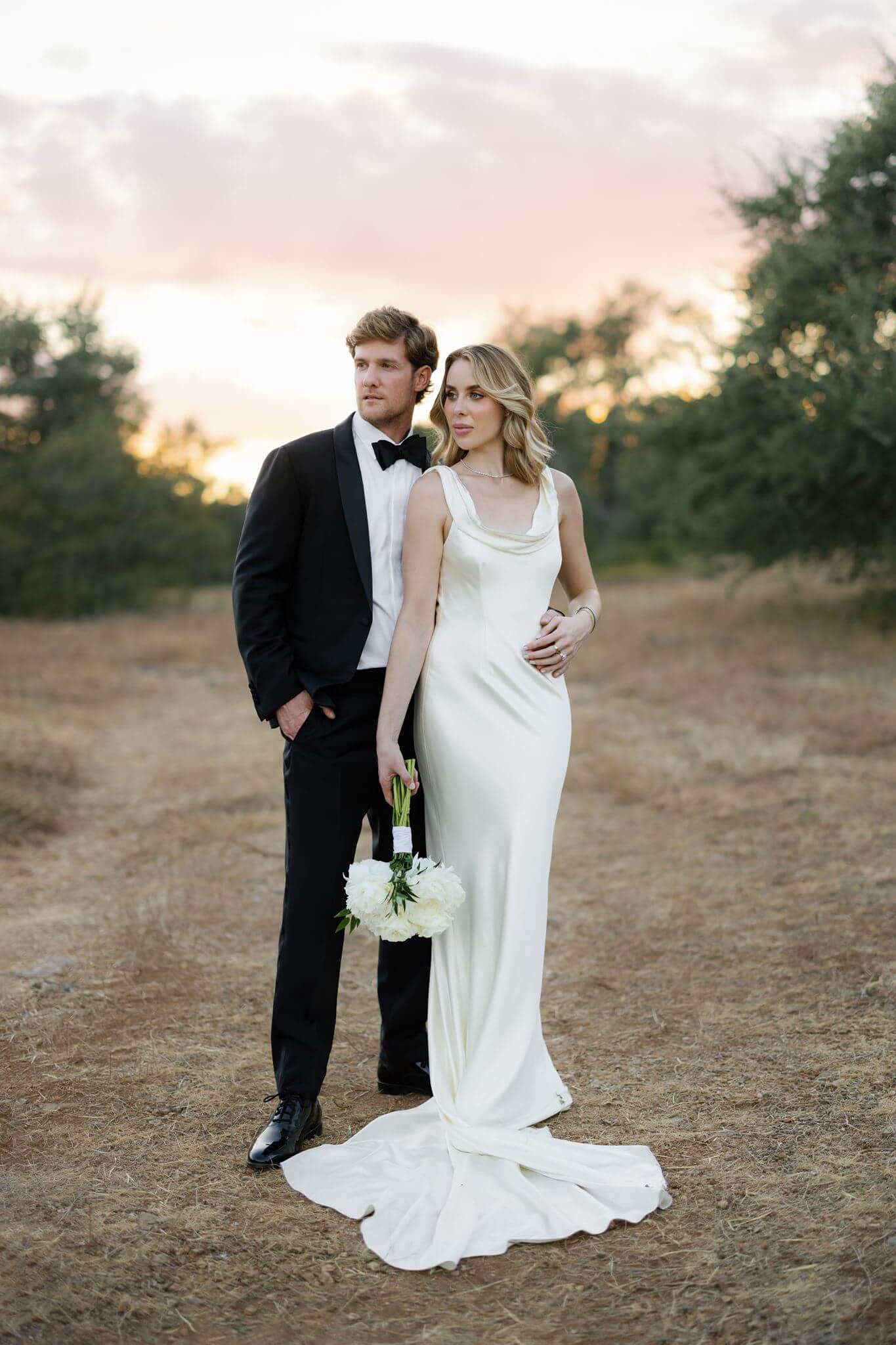 Bride and groom pose together in a dry grass field at sunset, with the bride holding a white flower bouquet.