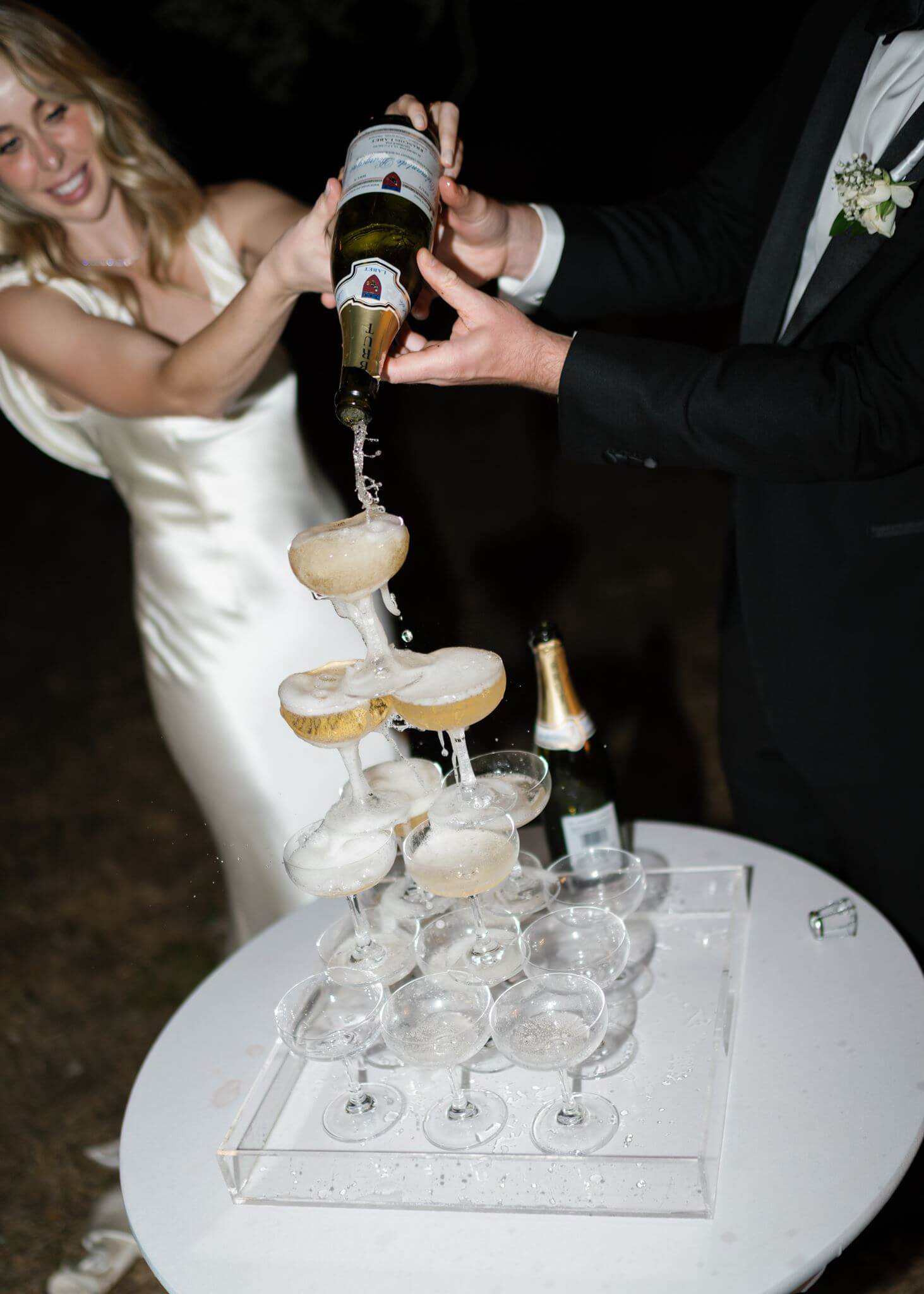 Bride and groom pour champagne over a tower of coupe glasses during their nighttime wedding reception.