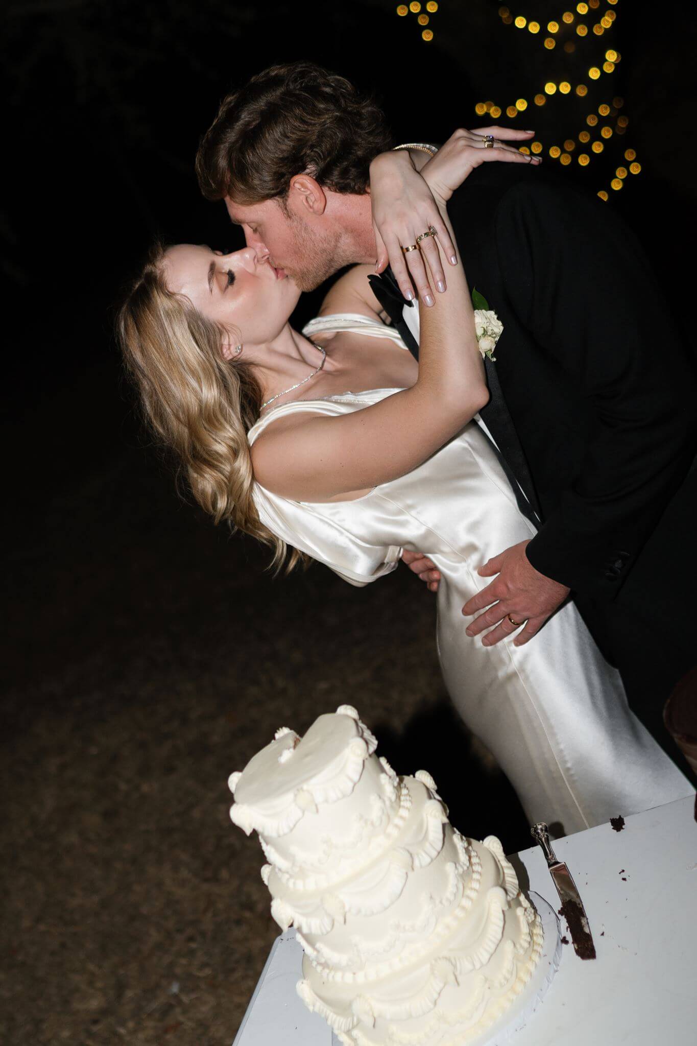 Newlyweds kiss beside their white wedding cake during the outdoor evening reception.