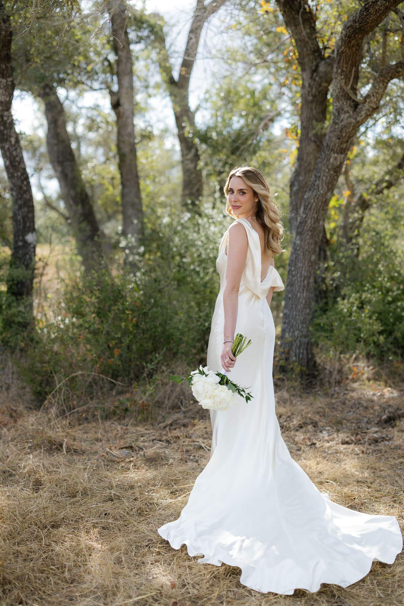 Bride stands among oak trees holding a white flower bouquet with the long train of her wedding dress behind her.