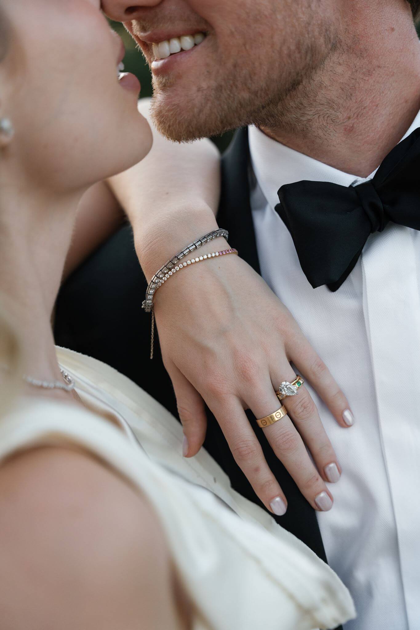 Close-up of a bride wrapping her arm around a smiling groom, showing her wedding rings and pearl bracelet.