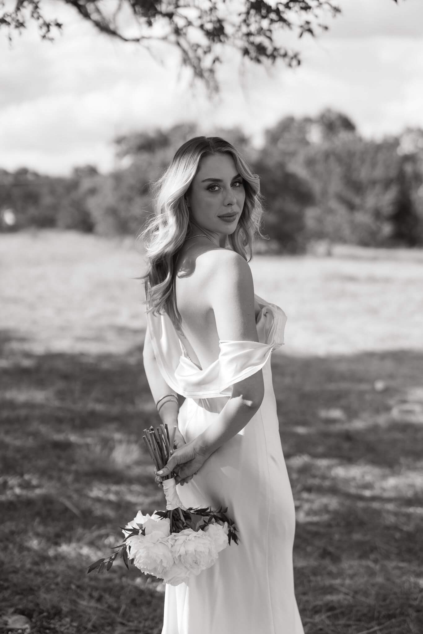 Black-and-white portrait of a bride looking over her shoulder in a field while holding her bouquet behind her back.