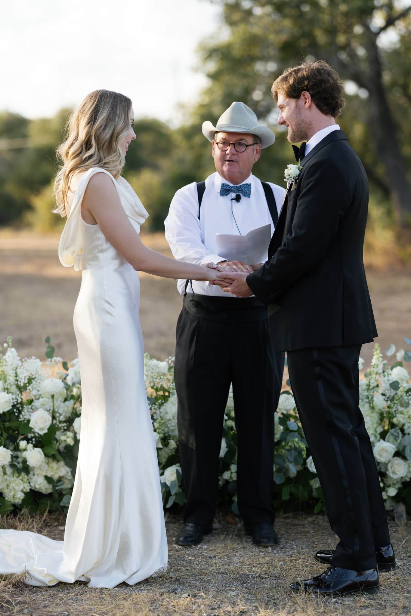 Bride and groom hold hands during their outdoor wedding ceremony with their officiant standing between them.