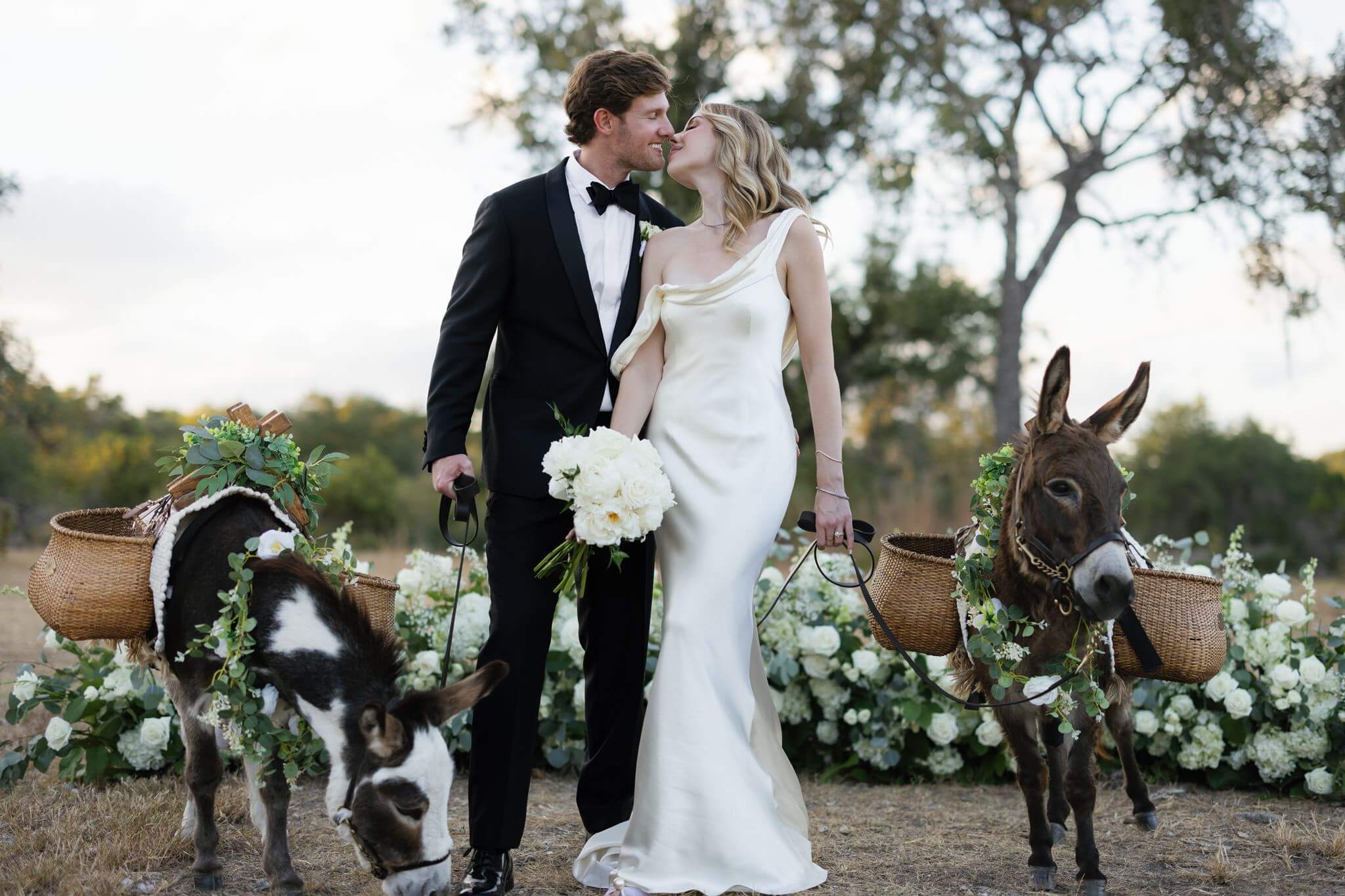 Newlyweds kiss while standing between two flower-adorned donkeys at their outdoor wedding.