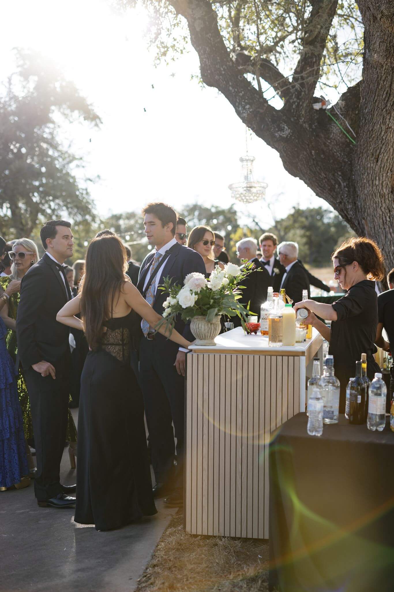 Wedding guests gather around an outdoor bar beneath a large tree as sunlight filters through the reception space.