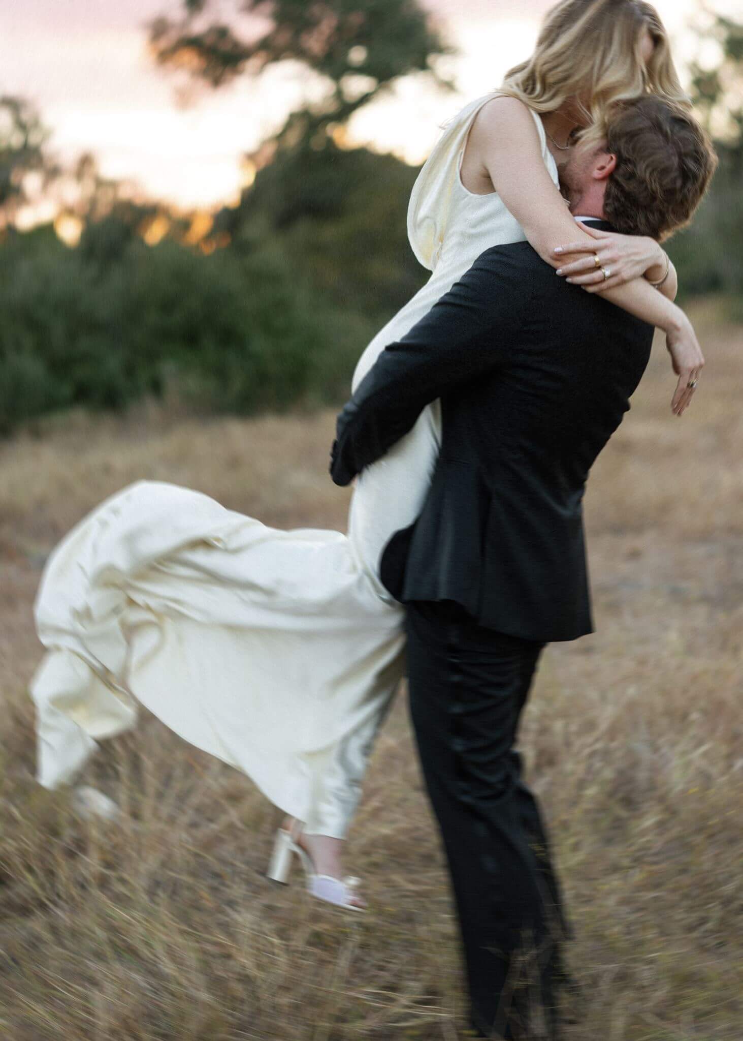 Groom lifts the bride into his arms in a grassy field as her satin wedding dress flows around them.
