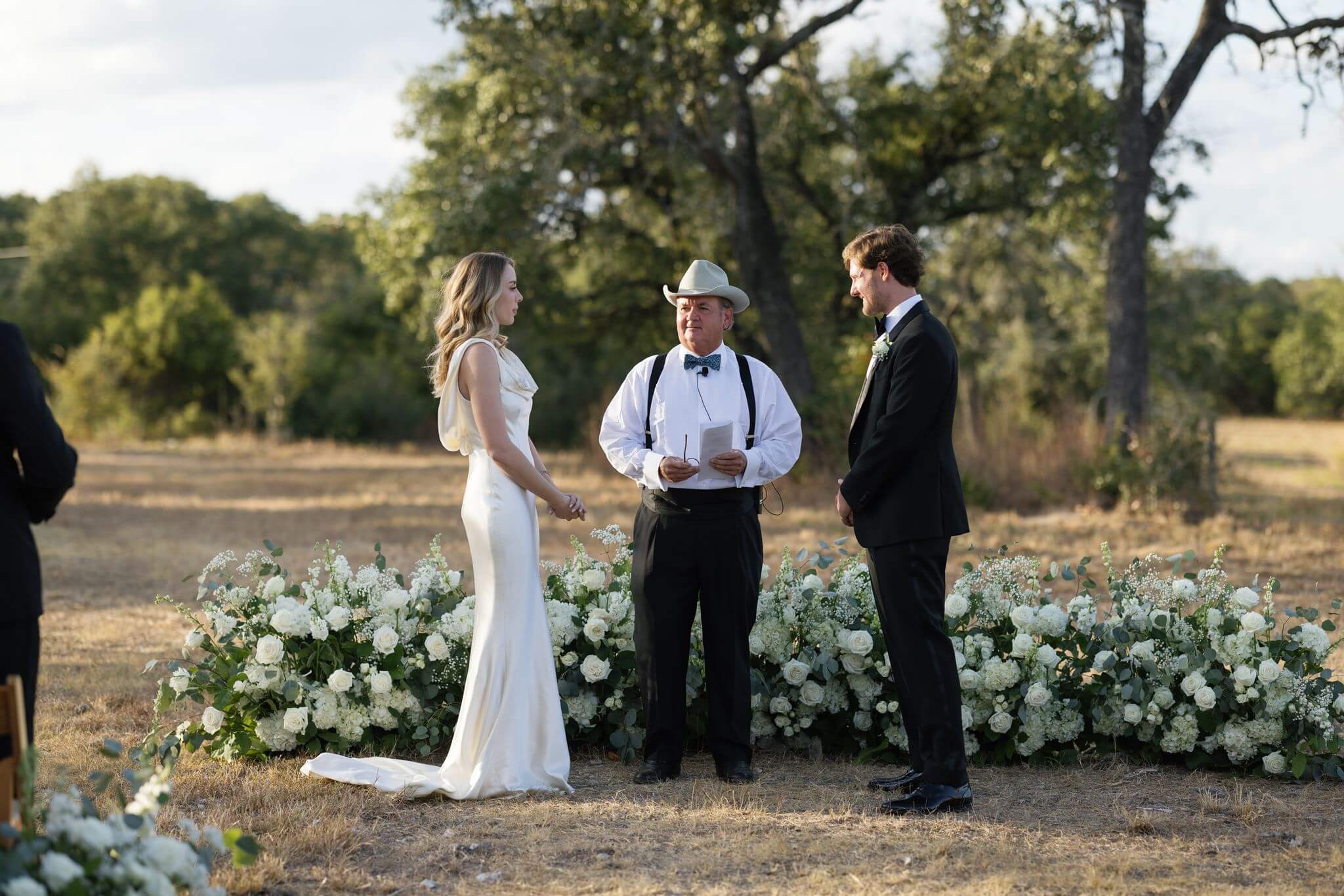 Wide view of a bride and groom standing with their officiant at an outdoor ceremony framed by low white floral arrangements.