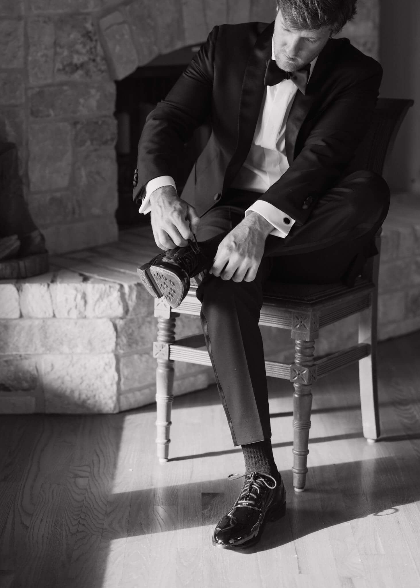 Black-and-white photo of a groom in a tuxedo tying his dress shoe while seated beside a stone fireplace.