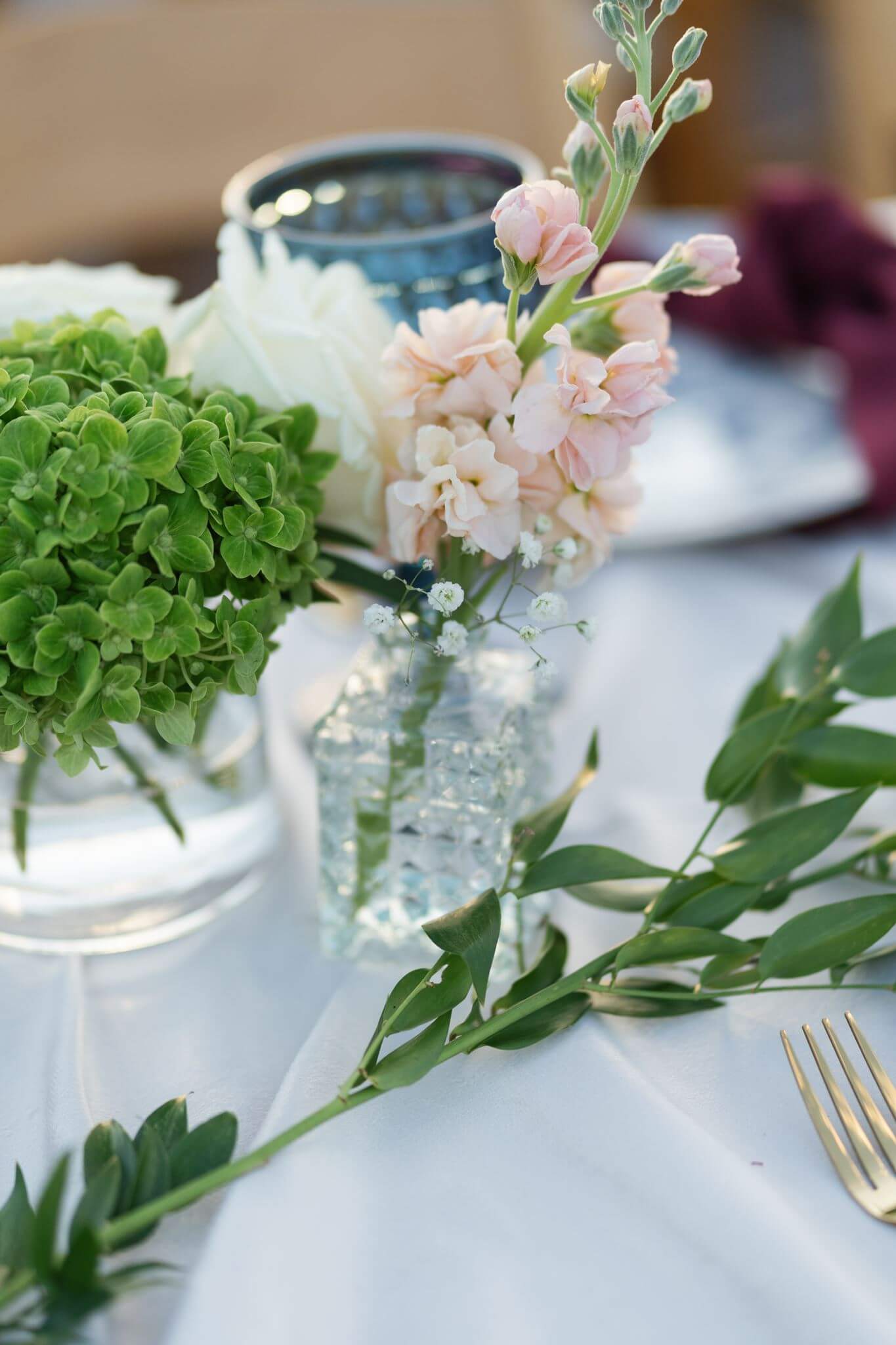 Close-up of a wedding table arrangement with green hydrangeas, pale pink flowers, greenery, and textured glassware.