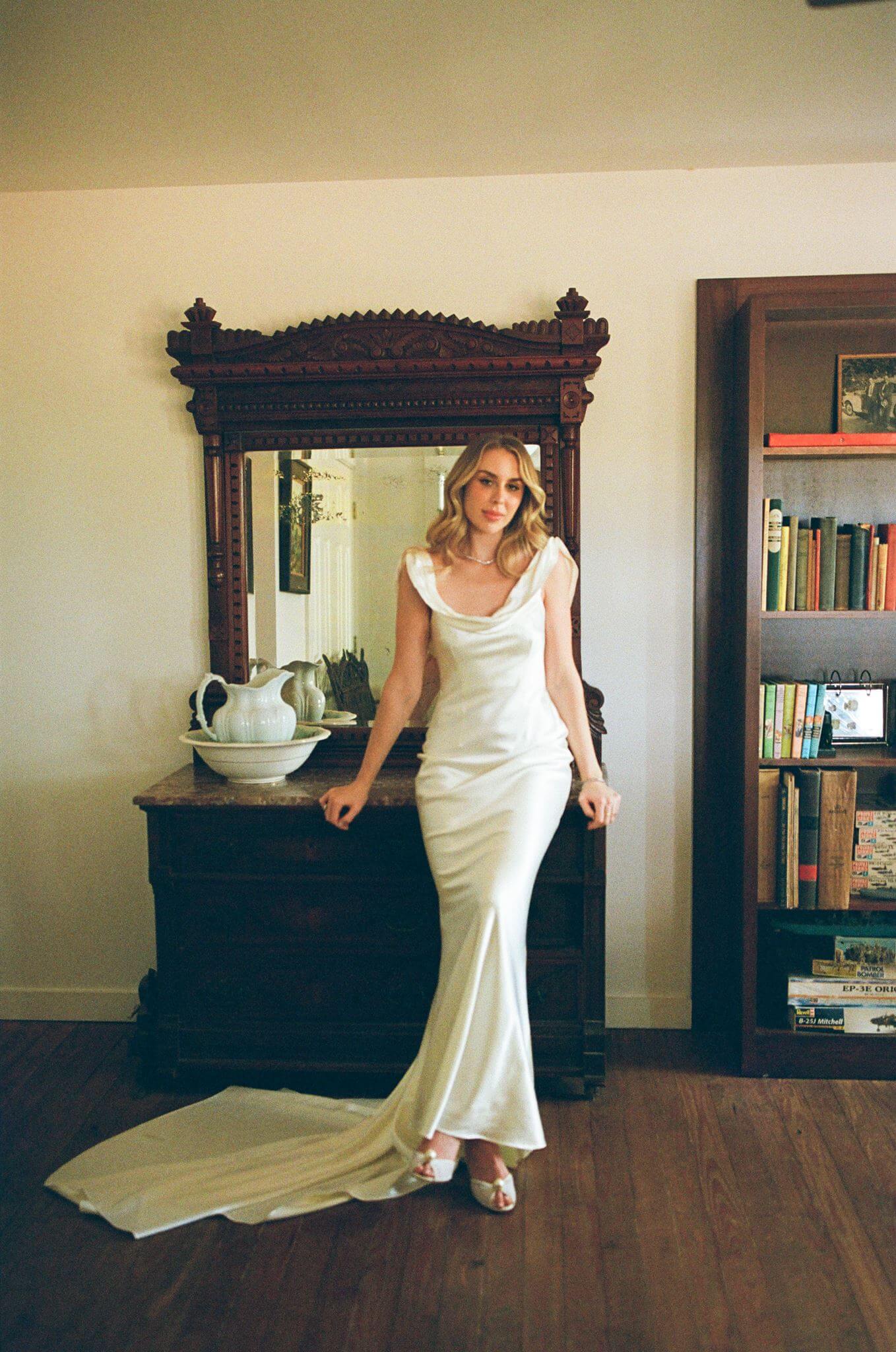Film-style bridal portrait of a bride in a satin wedding dress leaning against an antique dresser inside a bedroom.