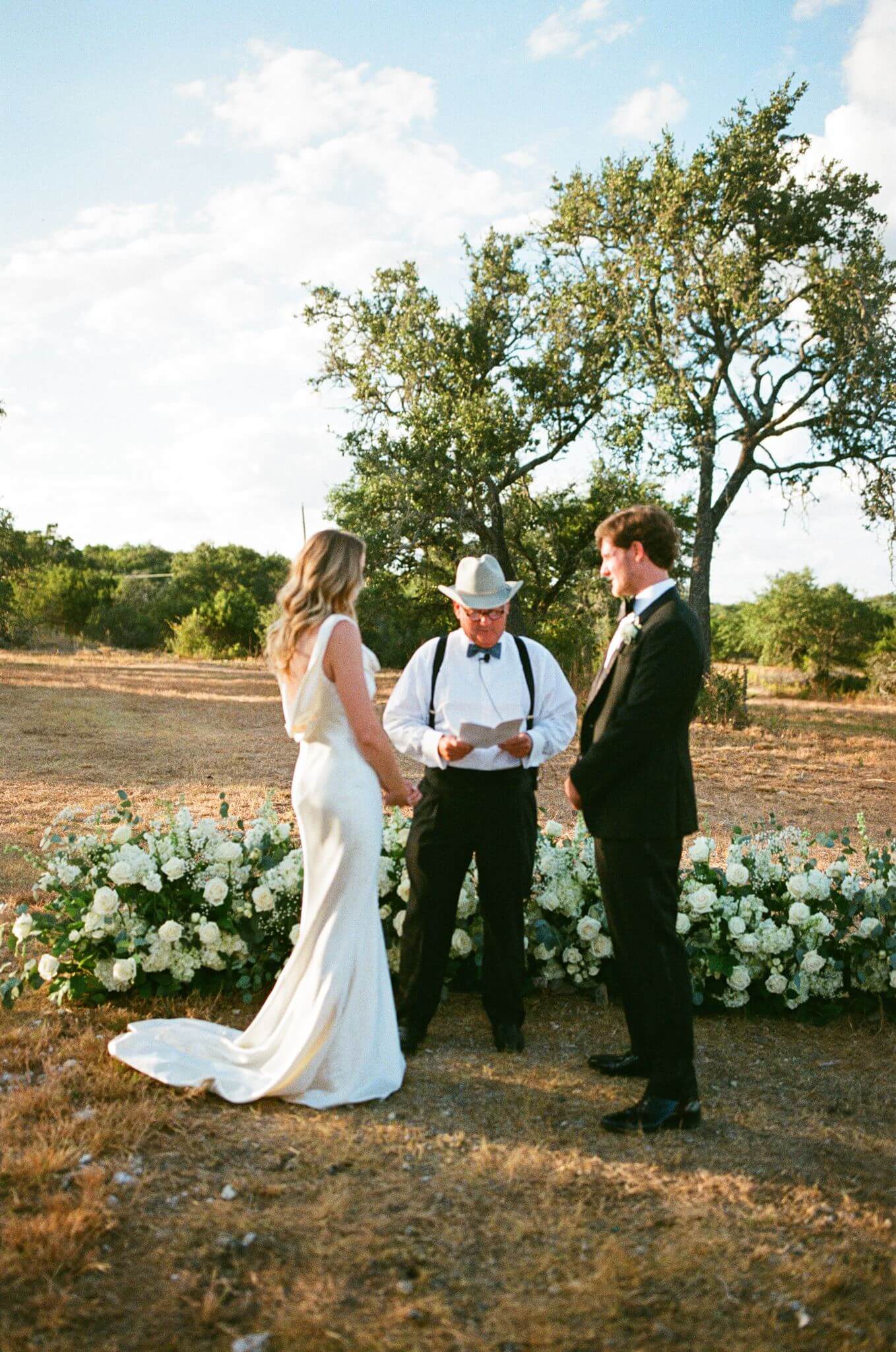 Bride and groom stand with their cowboy-hat-wearing officiant during an outdoor ceremony surrounded by white flowers.
