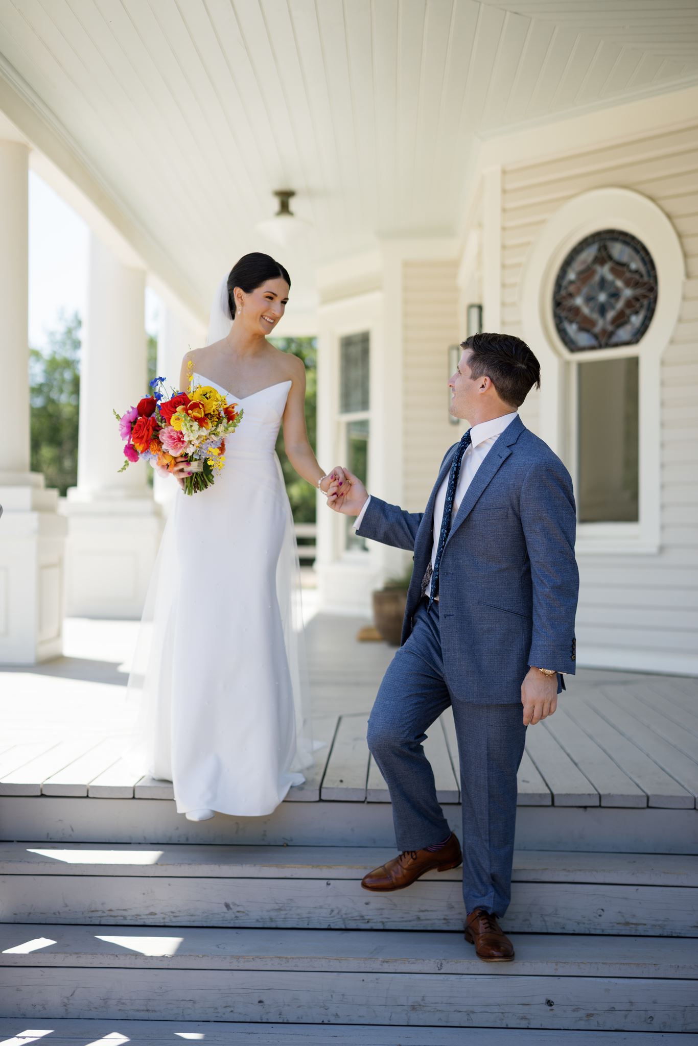 Bride standing on the porch steps while the groom kneels and holds her hand.