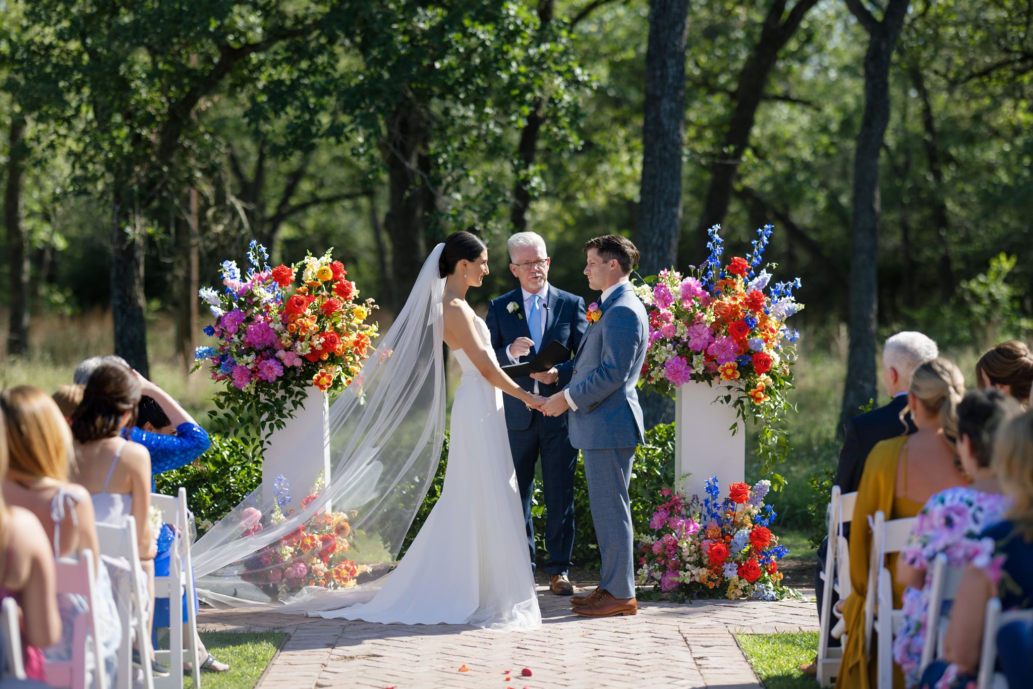 Bride and groom standing with their officiant during an outdoor ceremony framed by colorful flower arrangements.