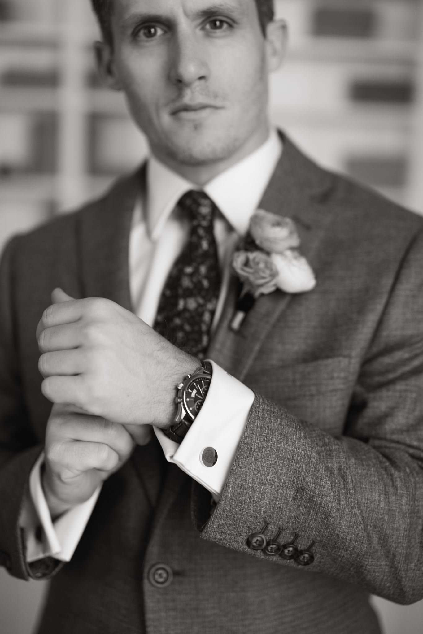 Black-and-white close-up of the groom adjusting his cuff while wearing a patterned tie and boutonniere.