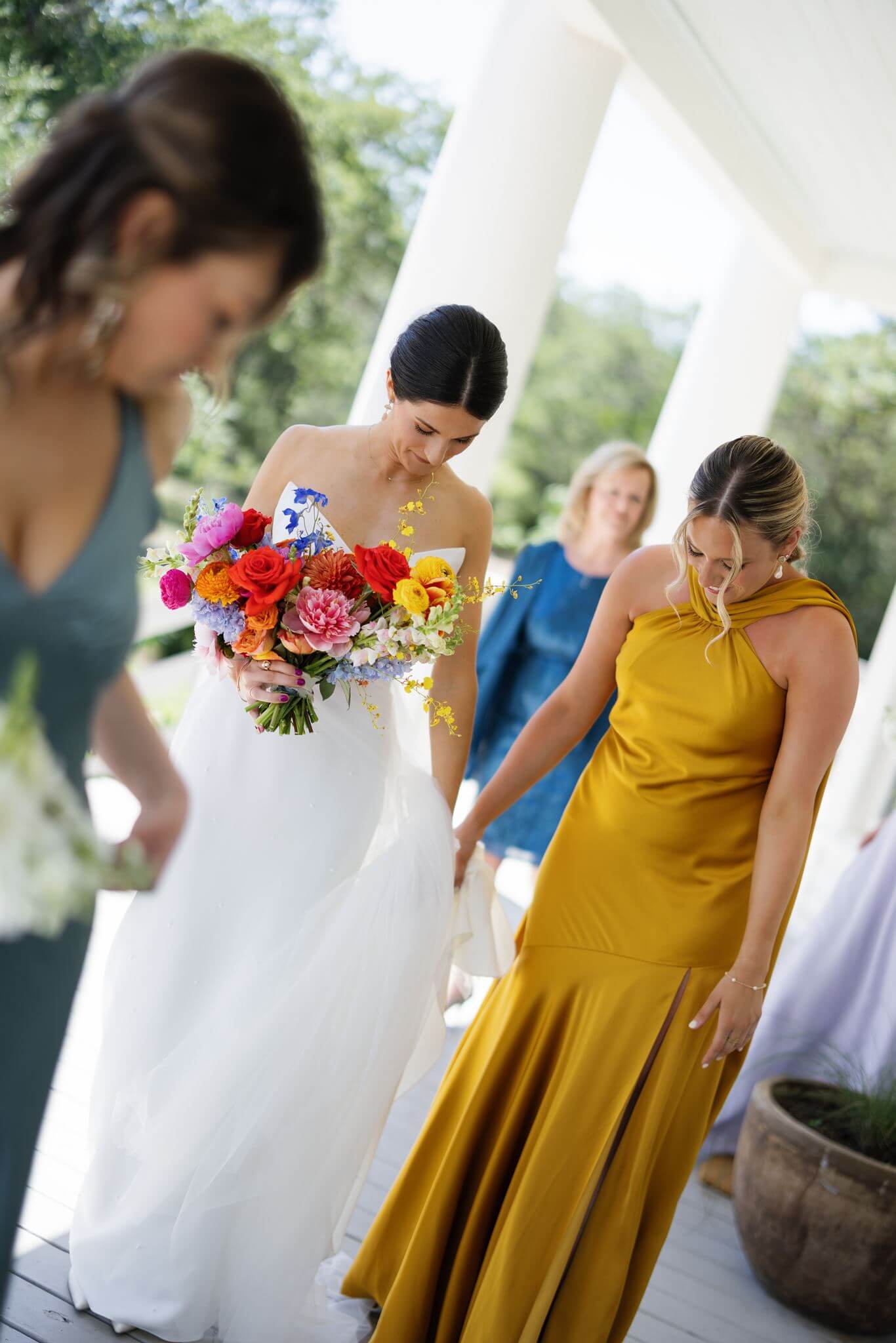 Bride holds her colorful wedding bouquet while a bridesmaid in a mustard yellow dress helps adjust her gown on the porch at The Grand Lady.