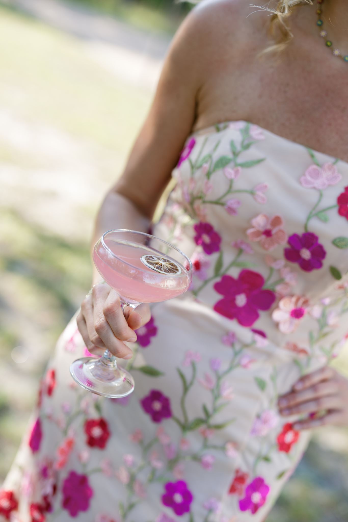 Wedding guest wearing a floral dress and holding a pink cocktail outdoors.