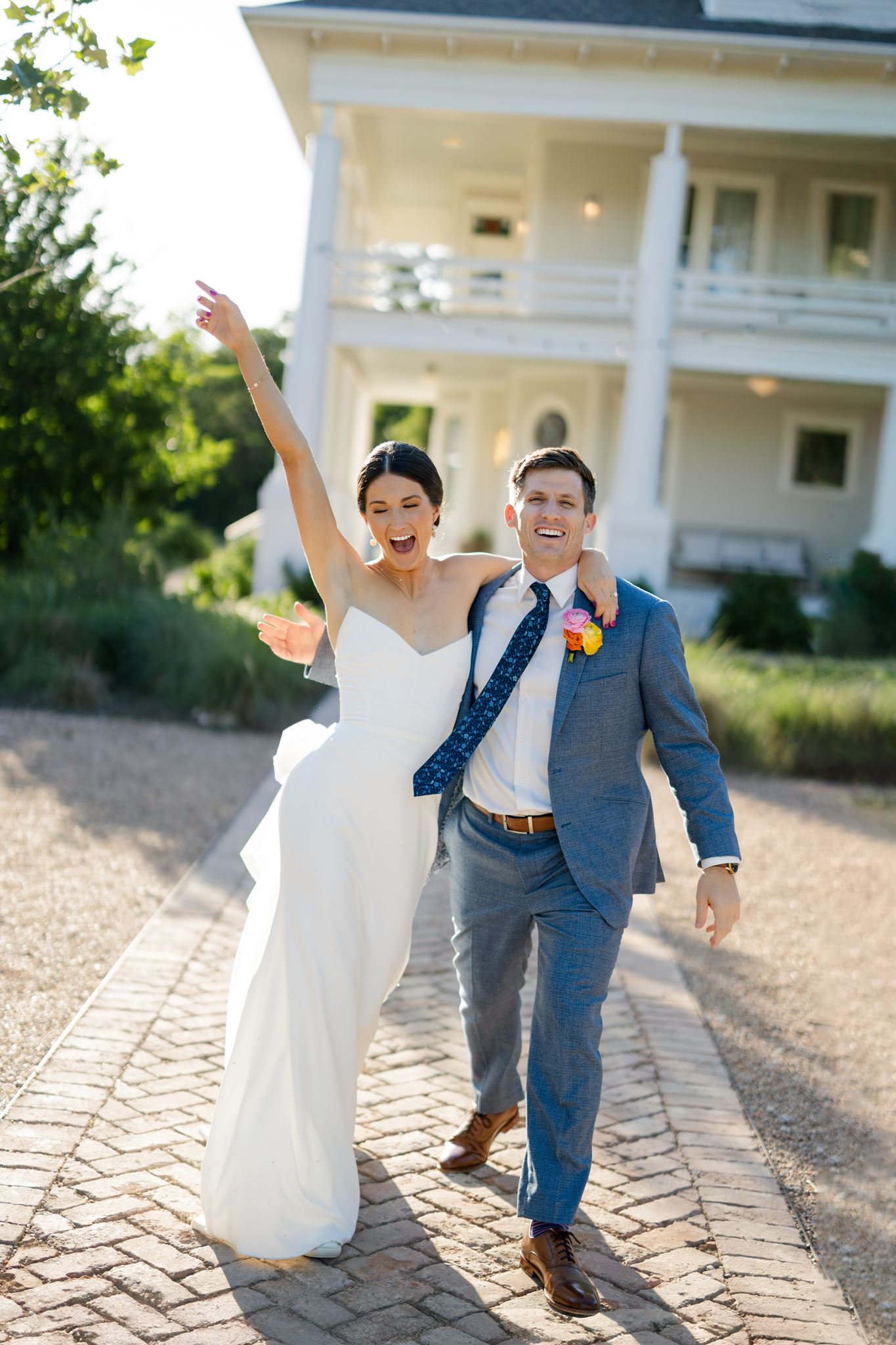Newlyweds smiling and raising their arms while walking together outside the white wedding venue.