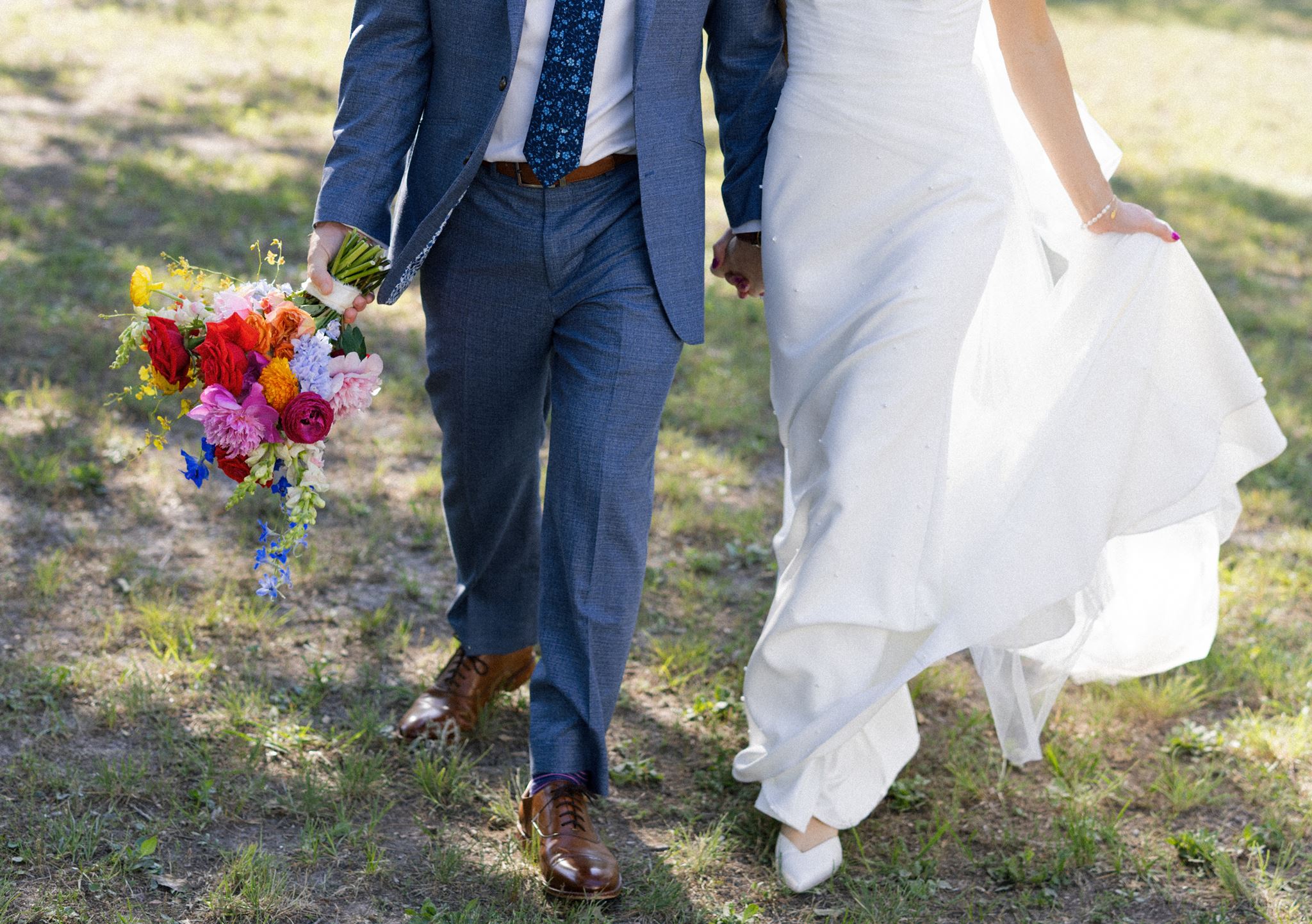 Bride and groom walking hand in hand outdoors with their bouquet between them.