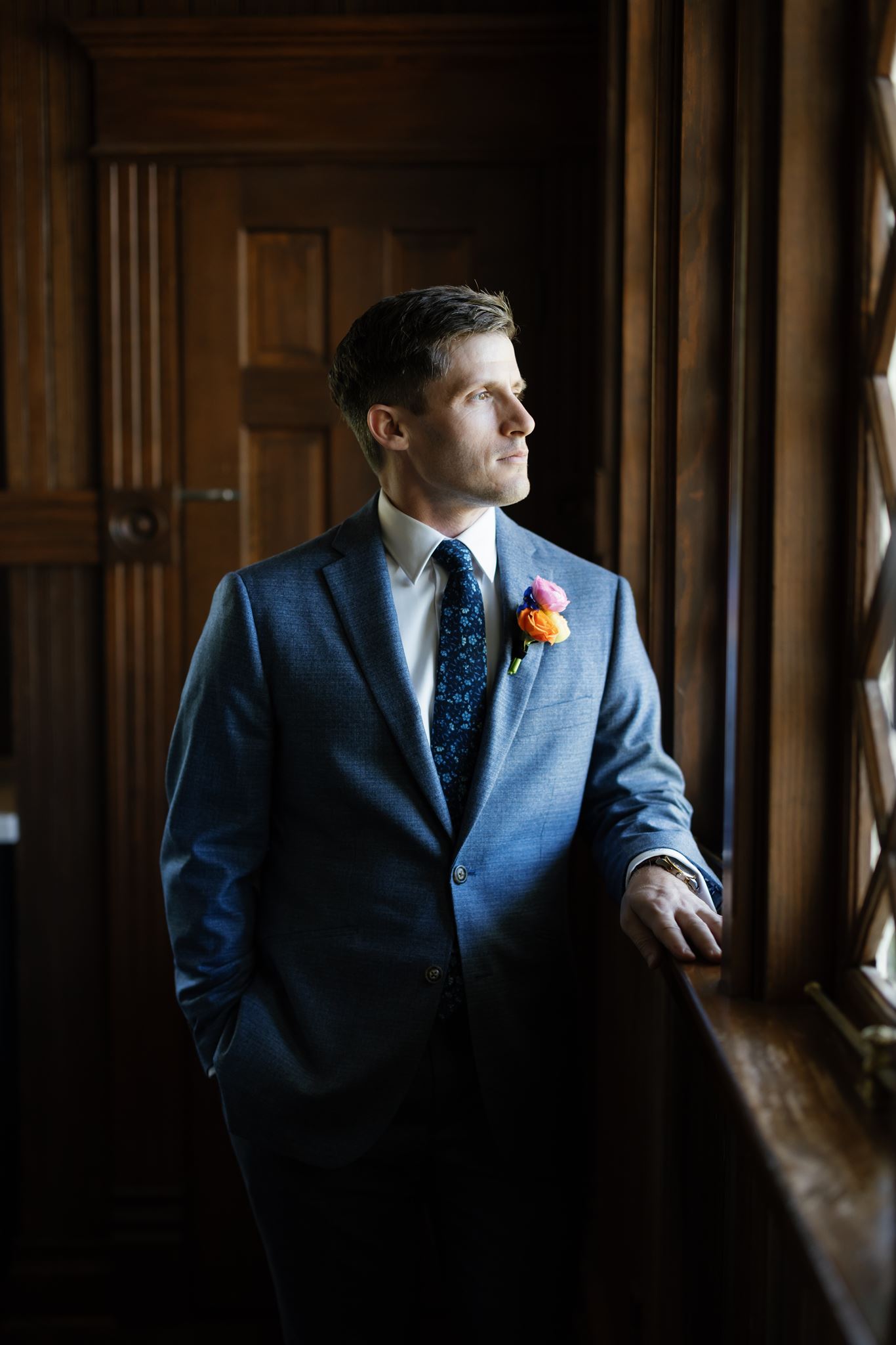 Groom in a blue suit standing beside a wooden staircase in a dark, wood-paneled room.