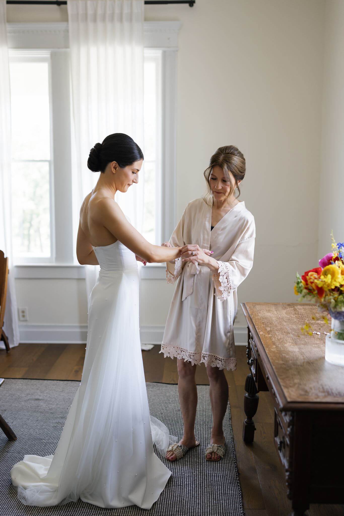 Bride getting ready in a bright room while a woman helps adjust her wedding dress.