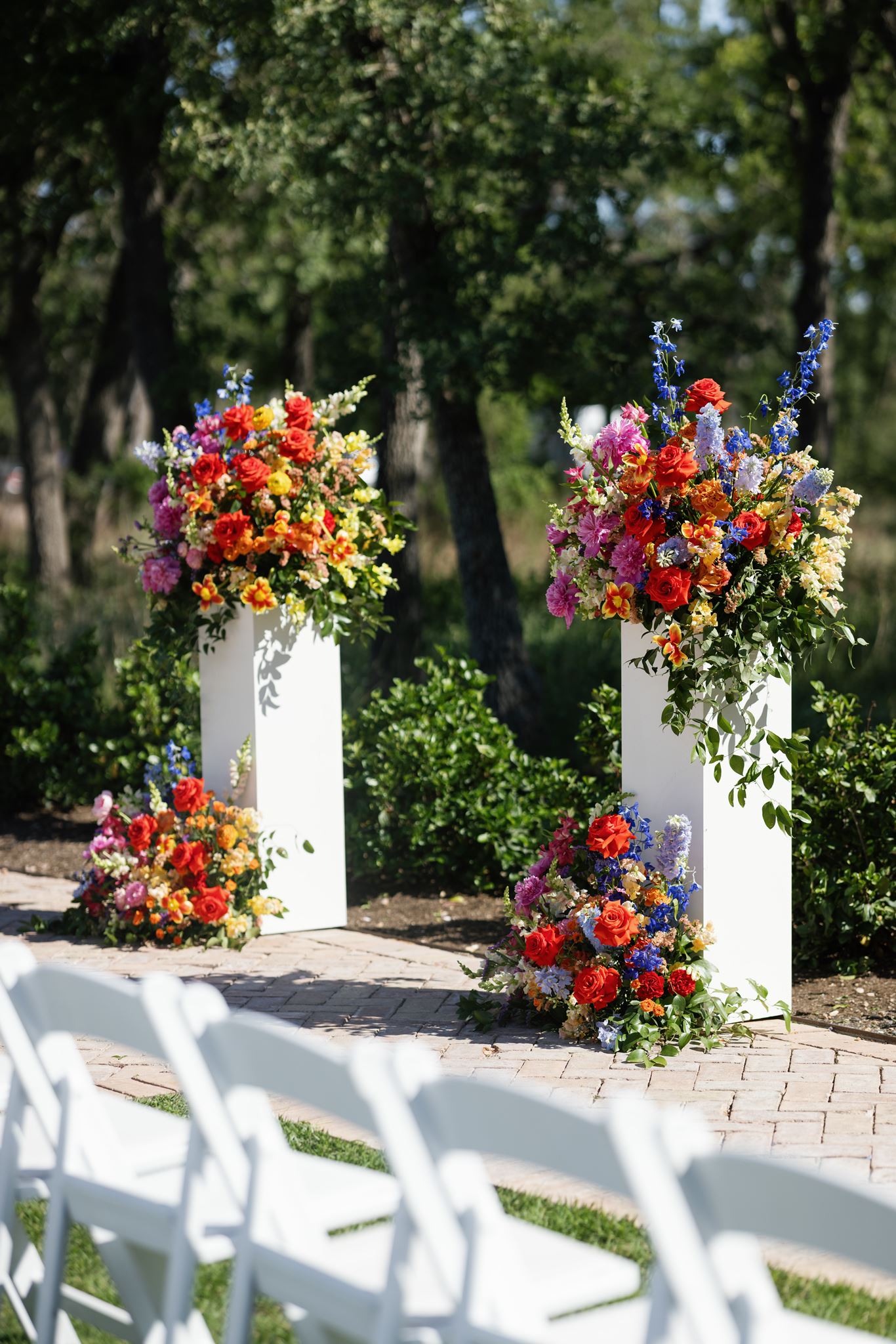Outdoor wedding ceremony setup with white chairs and two tall floral arrangements framing the aisle.
