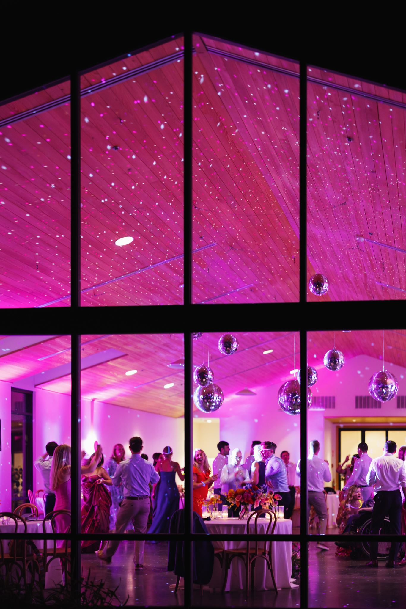 Wedding reception glowing with pink lighting as guests dance behind large glass windows.