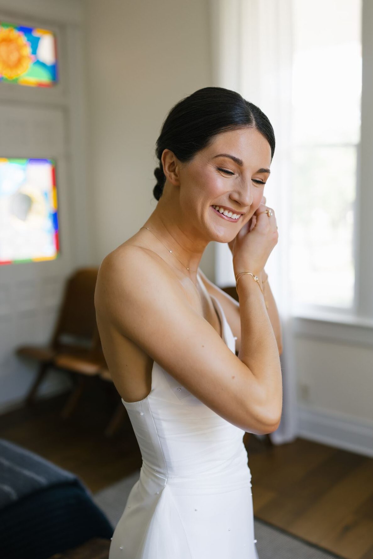 Bride smiling while putting on an earring in a sunlit room before the wedding.