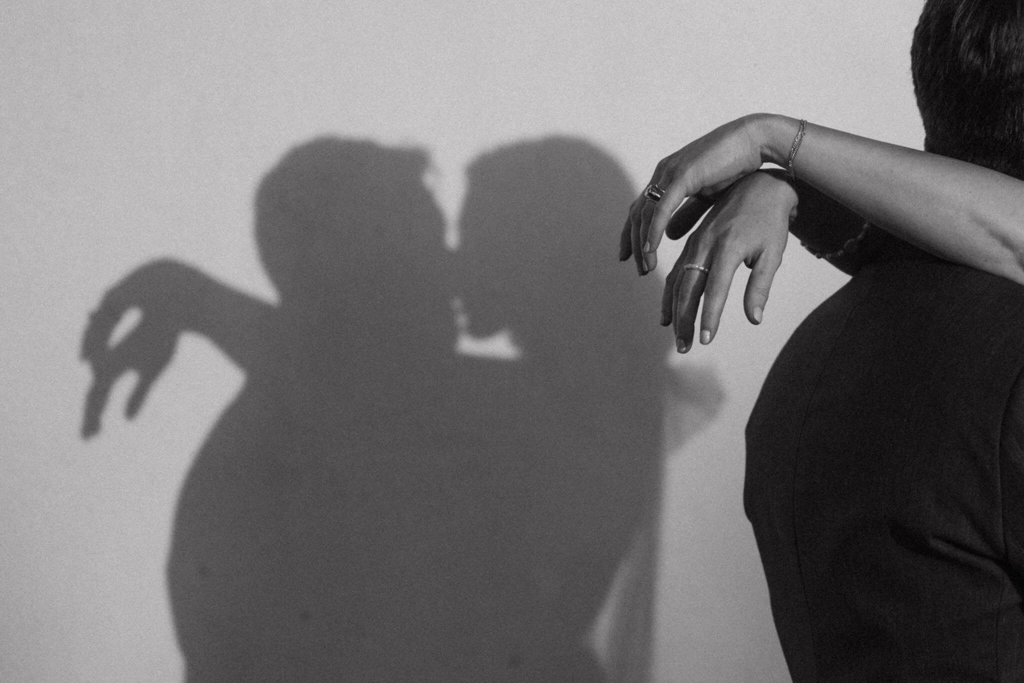 Black-and-white photograph of a couple's kissing silhouettes on a wall, with the bride's hand resting on the groom's shoulder in the foreground.