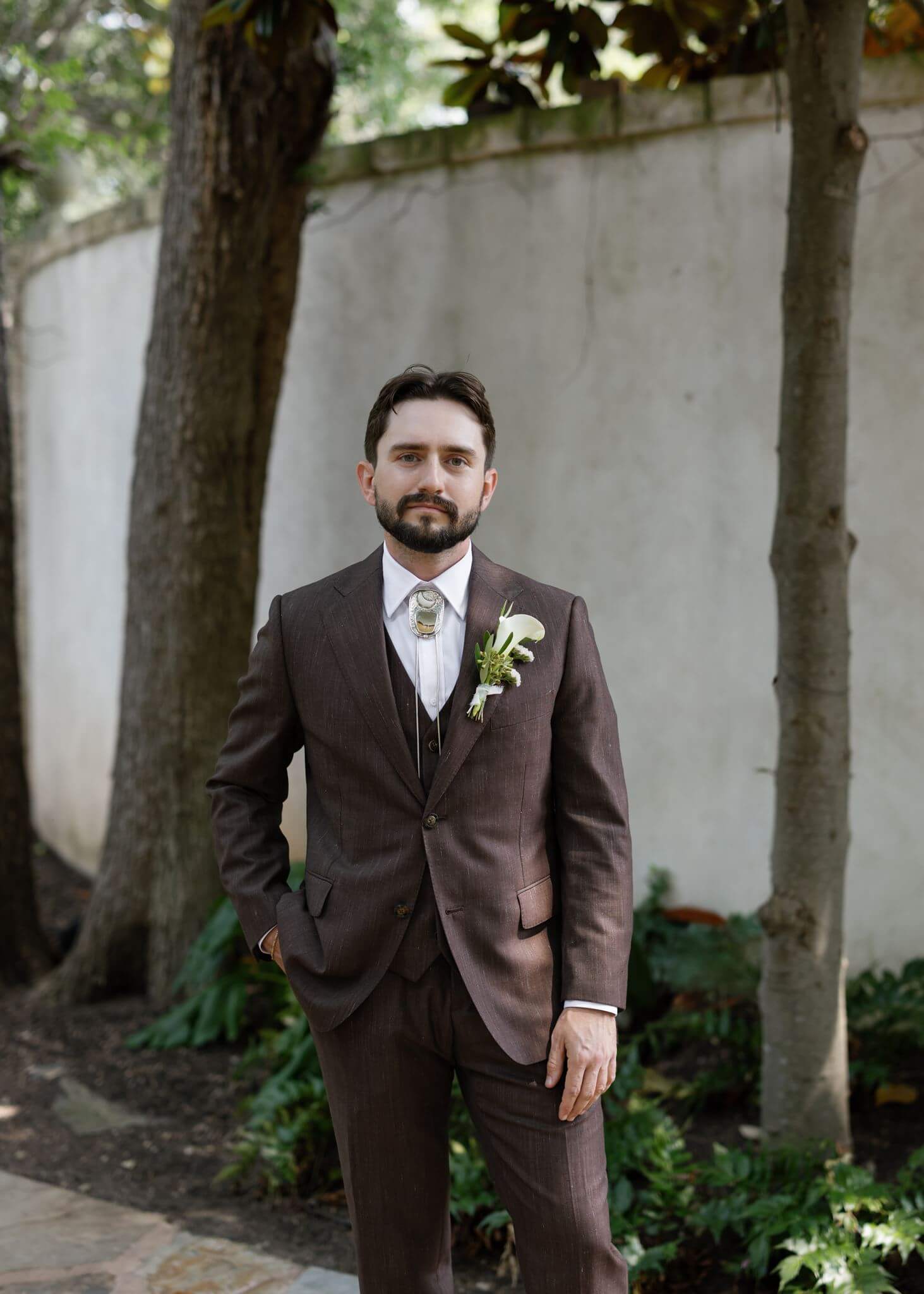 A groom wearing a dark brown suit and bolo tie poses along a garden pathway beneath the trees.