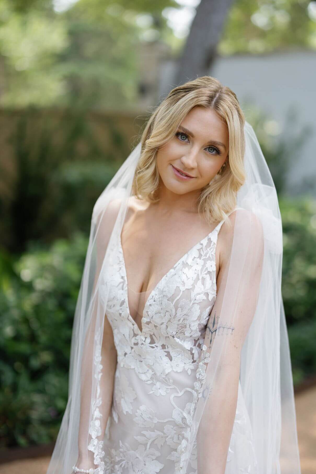 A bride poses alone in a floral lace wedding gown and long veil on a garden pathway surrounded by greenery.