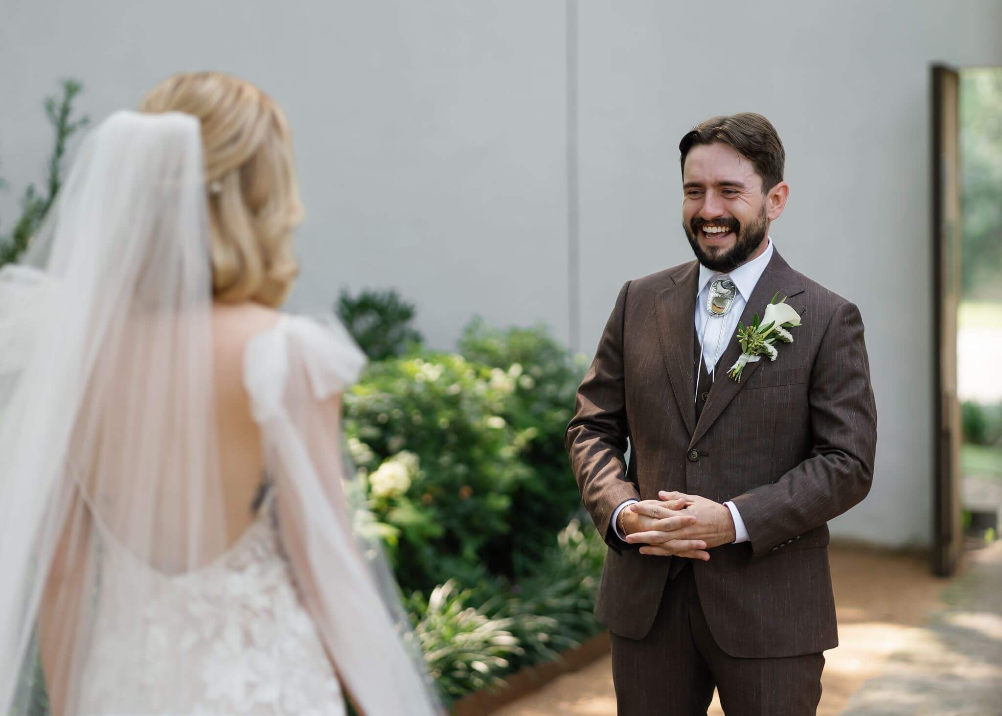 A groom smiles as he sees his bride for the first time during an outdoor first look surrounded by garden greenery.