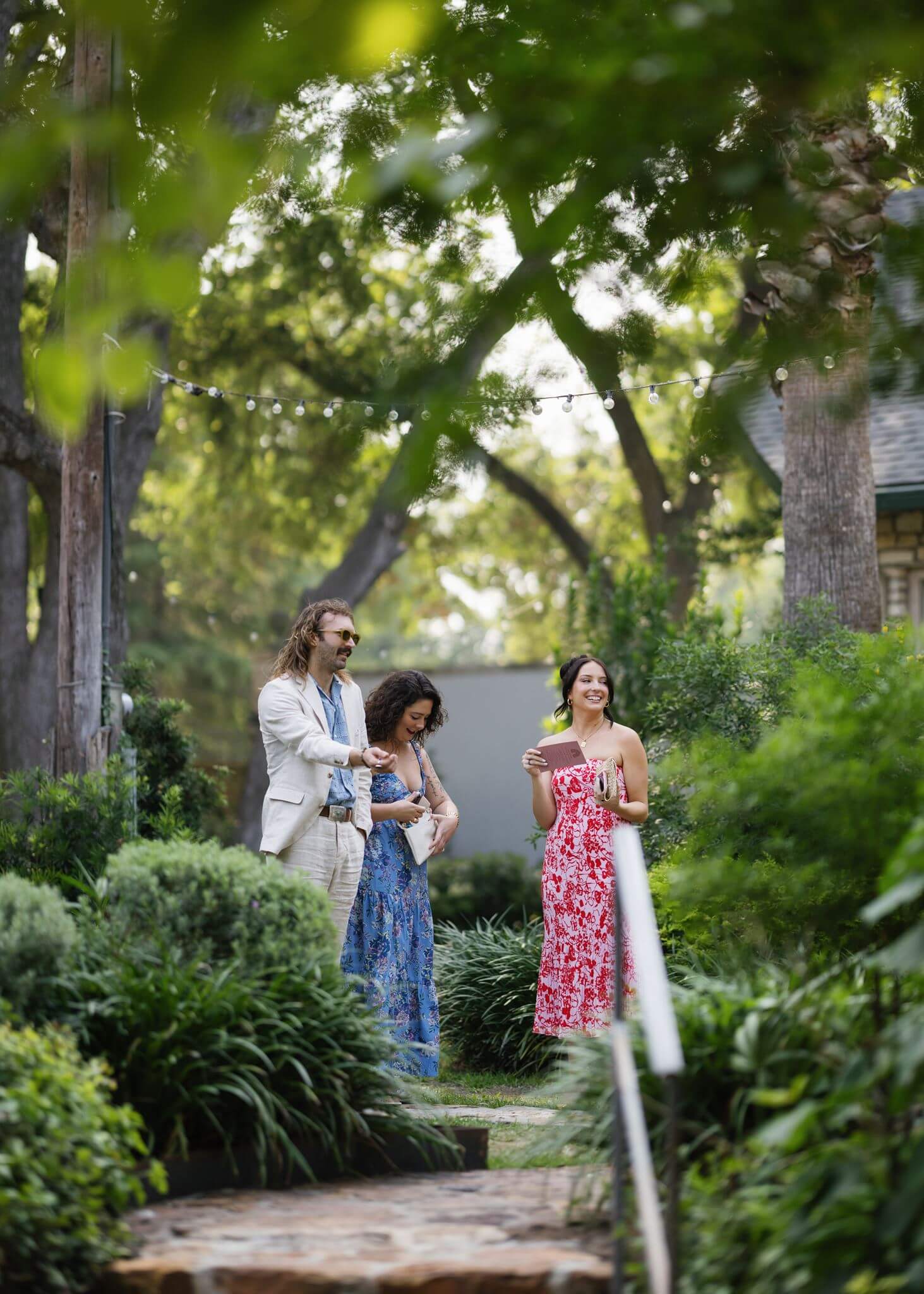Wedding guests wearing colorful dresses walk along a shaded garden path surrounded by lush greenery.