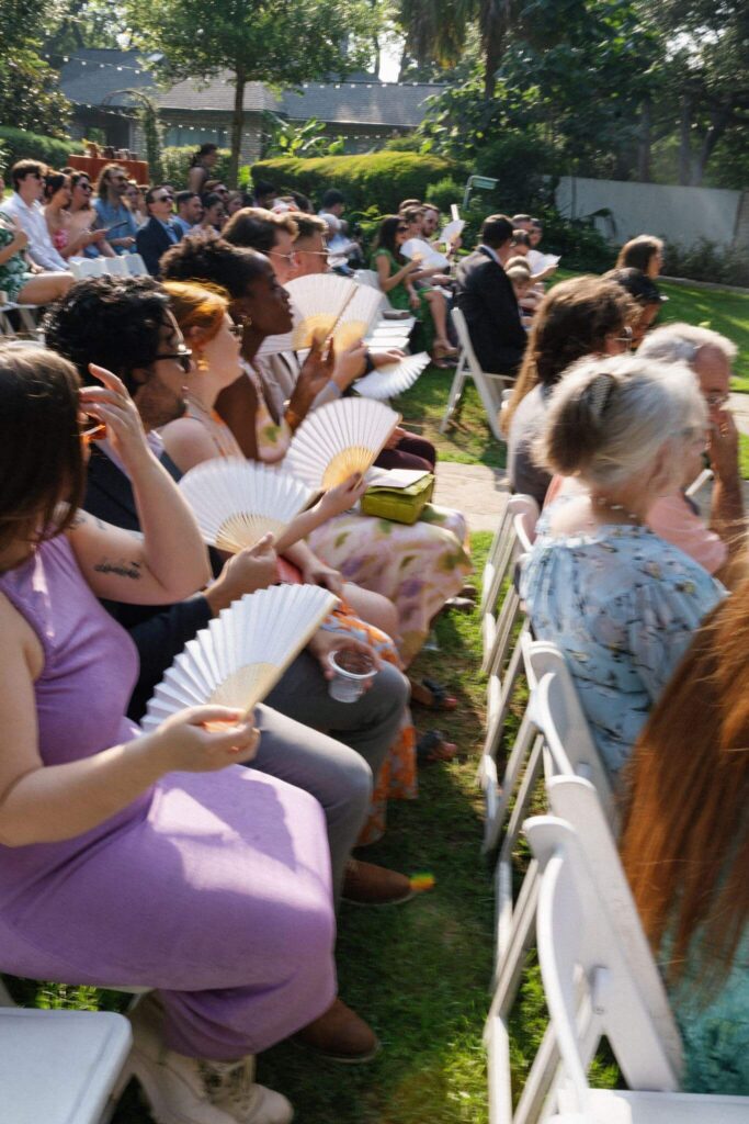 Wedding guests sit in rows of white chairs, cooling themselves with folding fans during an outdoor garden ceremony.