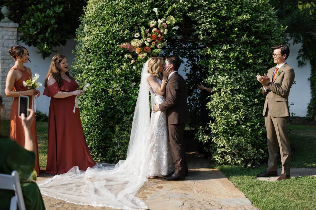 A bride and groom share their first kiss beneath a lush greenery arch during an outdoor wedding ceremony.