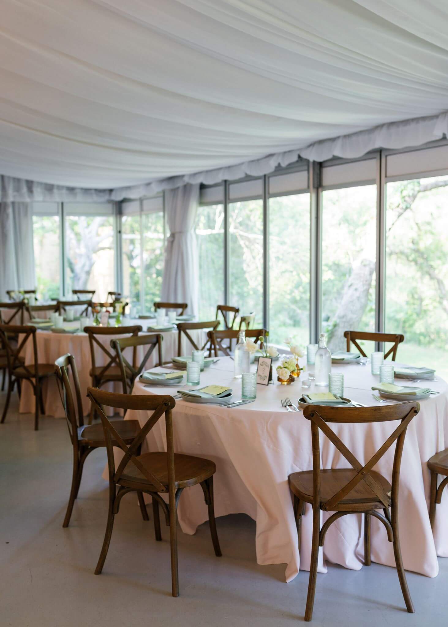 An indoor wedding reception space with round tables, pale pink linens, wooden cross-back chairs, and large windows overlooking the gardens.