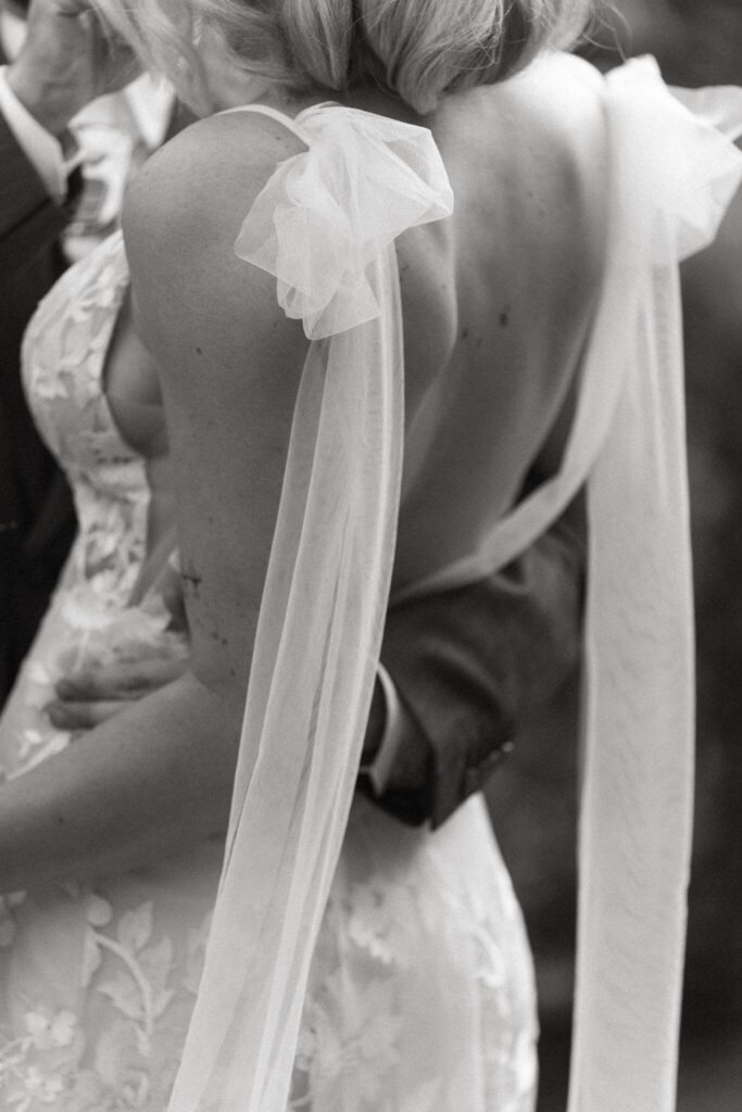 Black-and-white close-up of the groom's arm wrapped around the bride, highlighting the floral lace gown and sheer bow details on her veil.