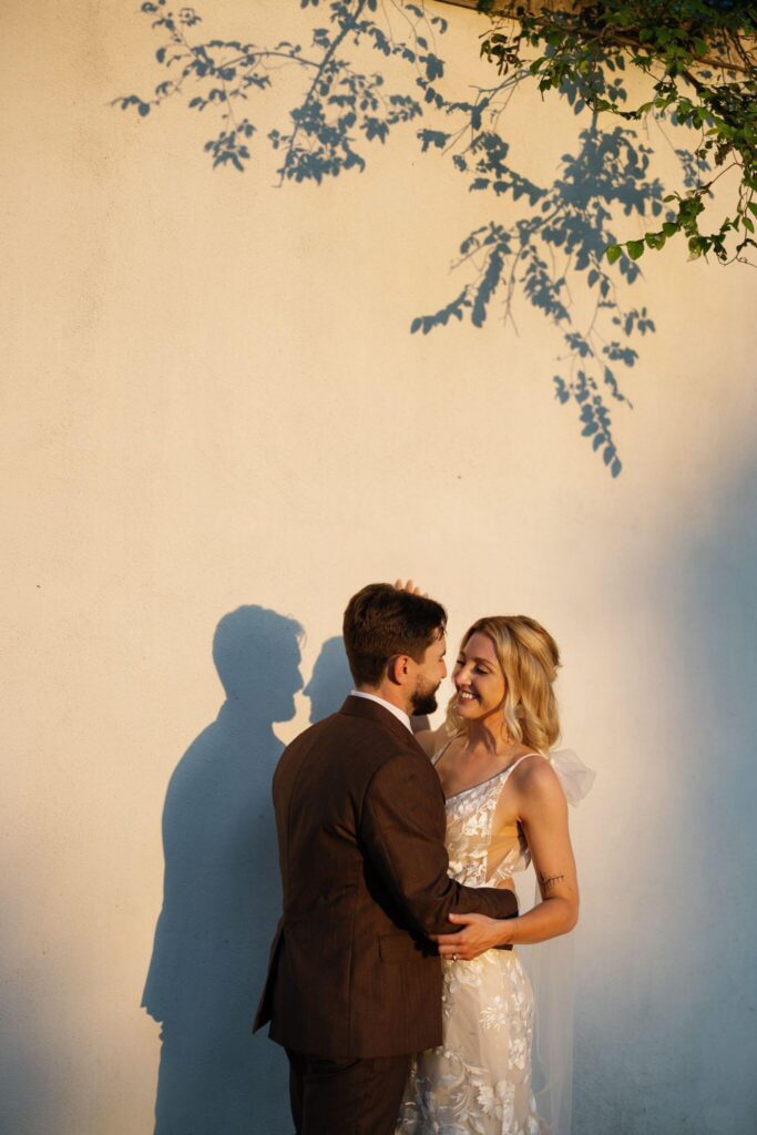 A bride and groom stand together against a pale wall in golden-hour sunlight, casting long shadows beside them.