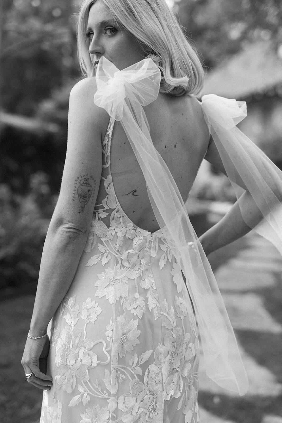 Black-and-white bridal portrait showing the open-back floral lace gown and sheer bow veil as the bride looks over her shoulder.